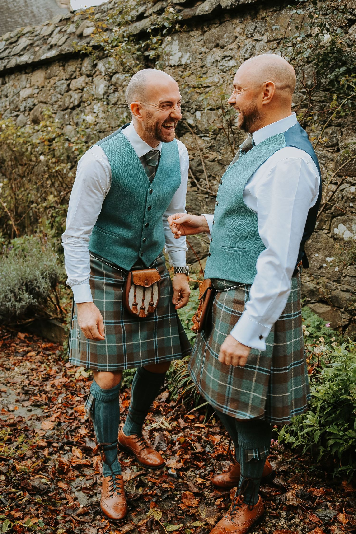 Two people in matching green vests and plaid kilts share a laugh while standing against a stone wall.