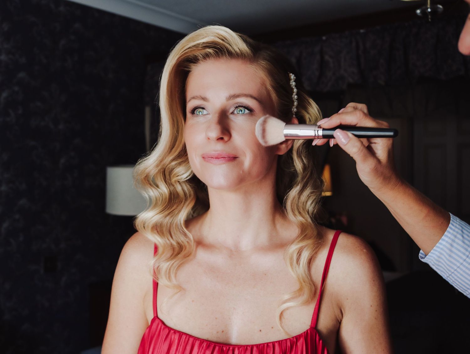 Makeup artist applies cosmetics with a brush to model wearing red spaghetti strap top.