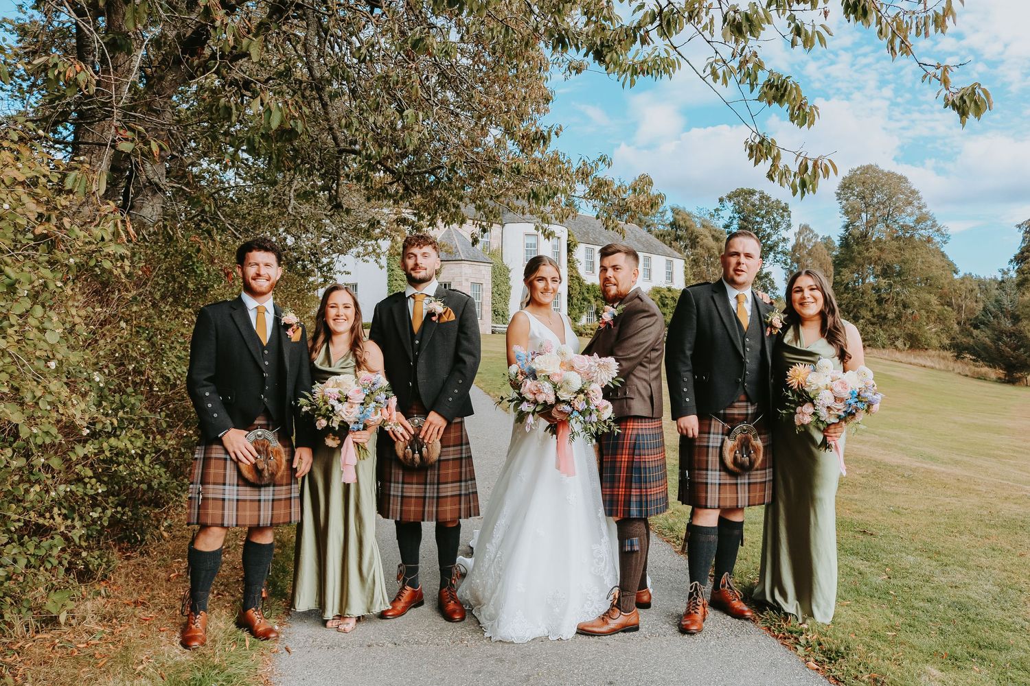 Wedding party in traditional Scottish kilts and formal attire poses together on sunny autumn day.