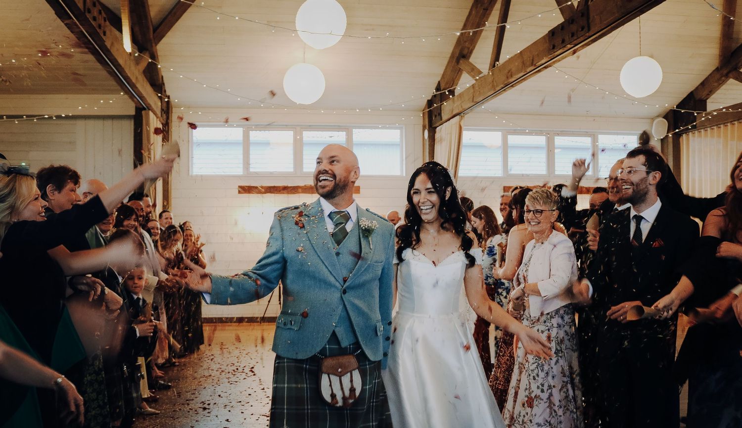 Wedding guests dance and celebrate together during a reception in a rustic barn venue with glowing pendant lights.