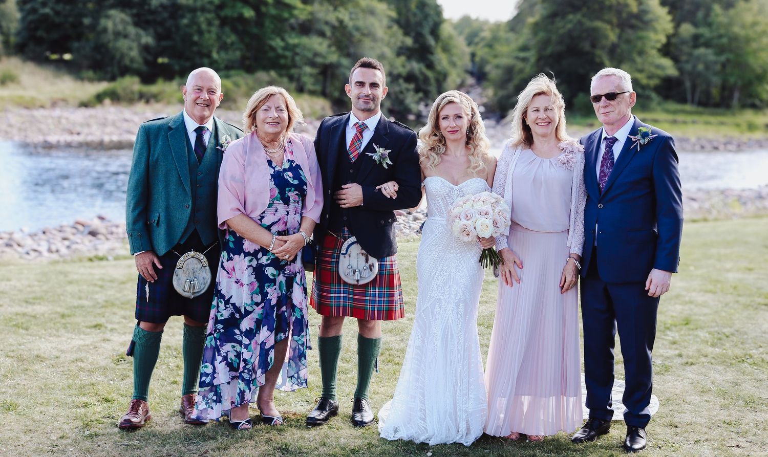 Group photo of guests in formal wear including kilts and evening dresses gathered outdoors by a waterside for a Scottish wedding.