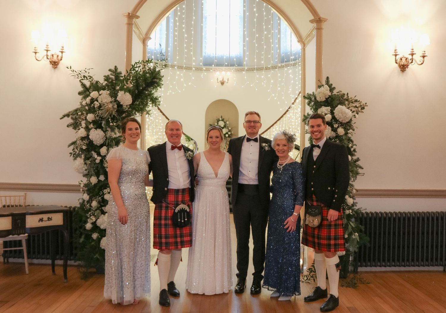 A wedding party poses under a floral arch with decorative wall sconces in an elegant indoor venue.