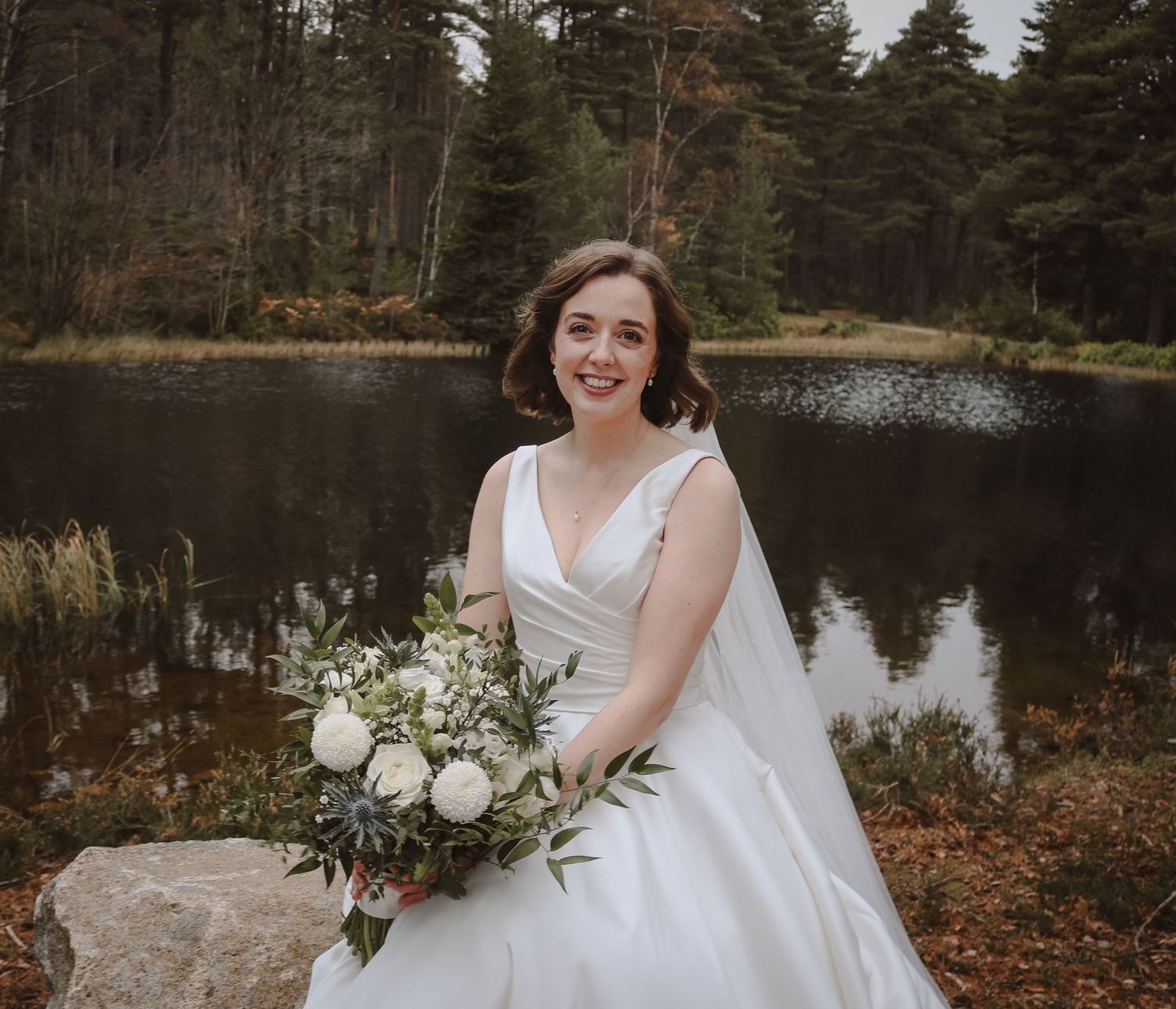 A bride in a white wedding dress sits on a rock beside a serene lake surrounded by evergreen trees.