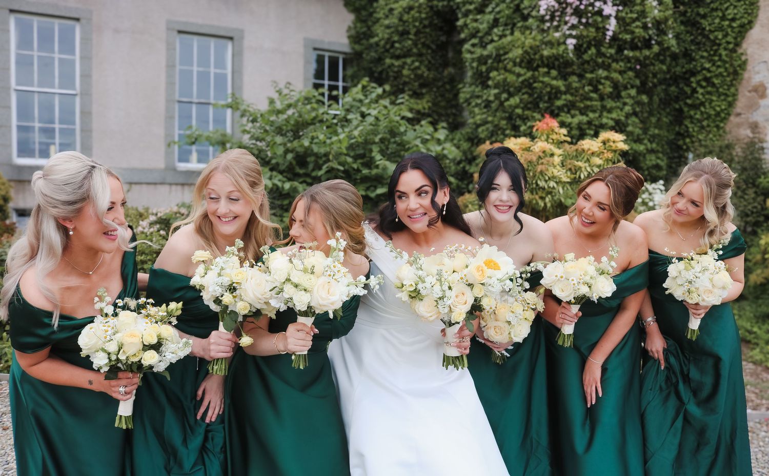 Bride in white gown with six bridesmaids in emerald green dresses holding white bouquets outside a stone venue.