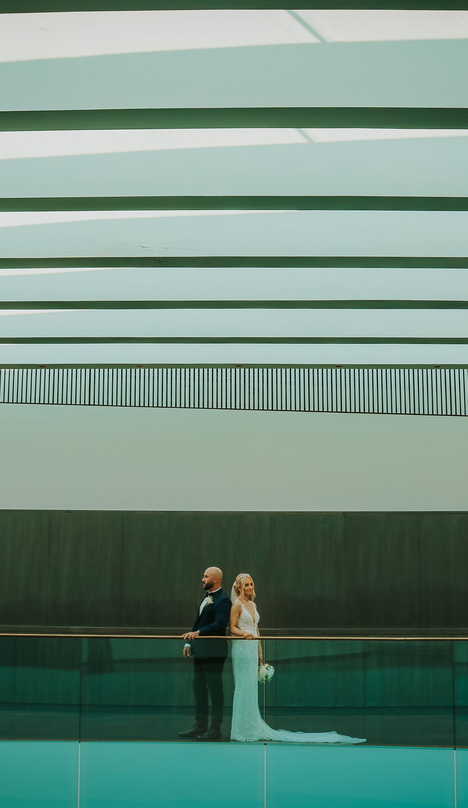 A couple in wedding attire stands back-to-back against a modern green architectural backdrop with geometric ceiling lines.