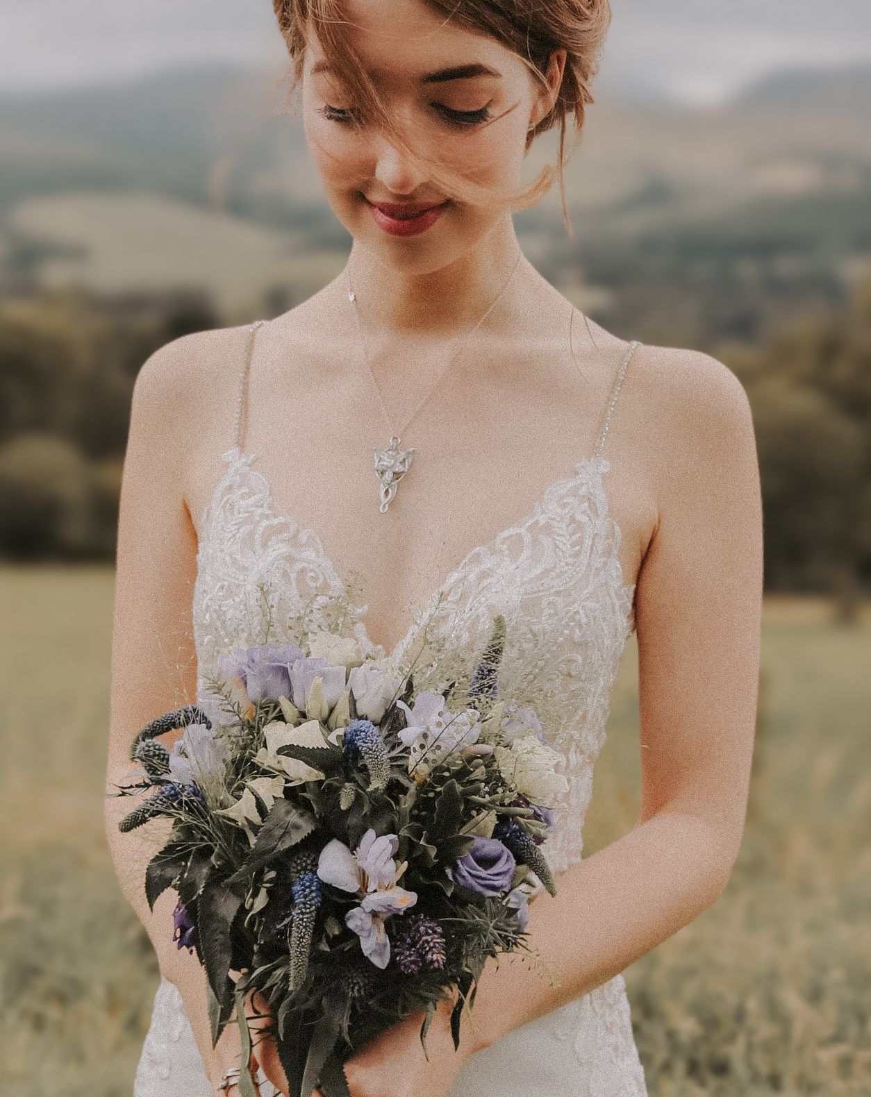 Bride in lace wedding dress holding delicate bouquet of purple and white flowers against mountainous backdrop.