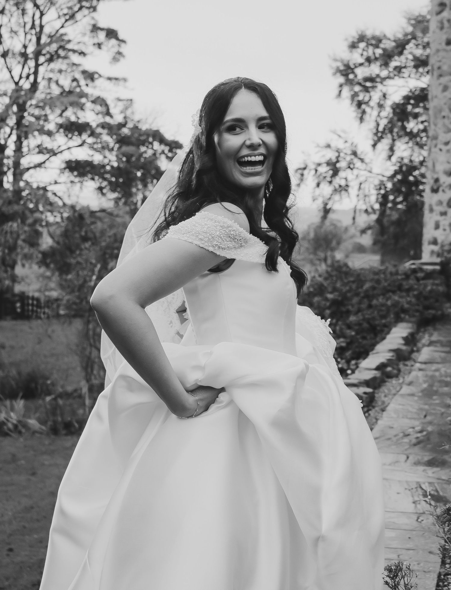 Joyful bride in a white lace wedding dress laughs while standing outdoors in a garden setting.