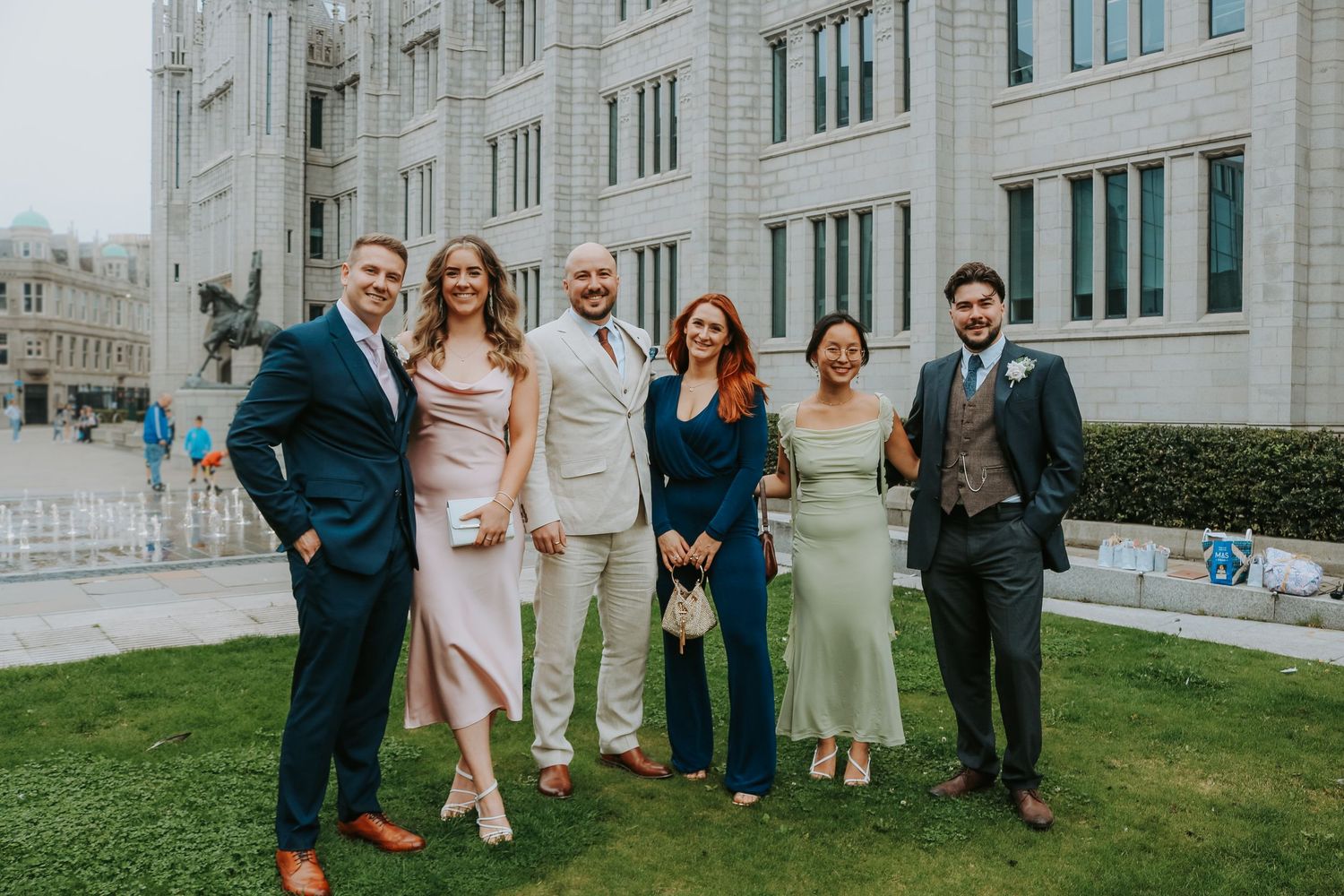 Group of wedding attendees pose together on grass outside elegant stone building in formal attire.