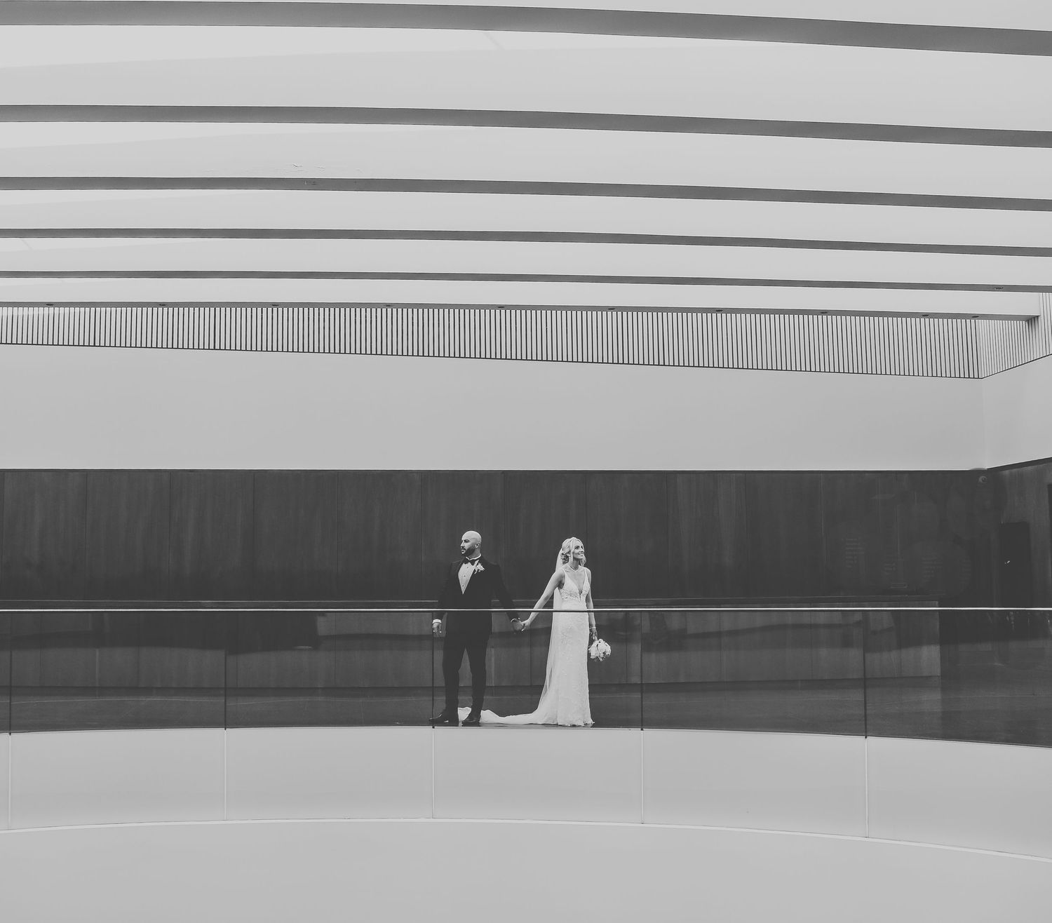 A couple in formal attire stand silhouetted against large curved architectural lines in a modern monochromatic interior space.