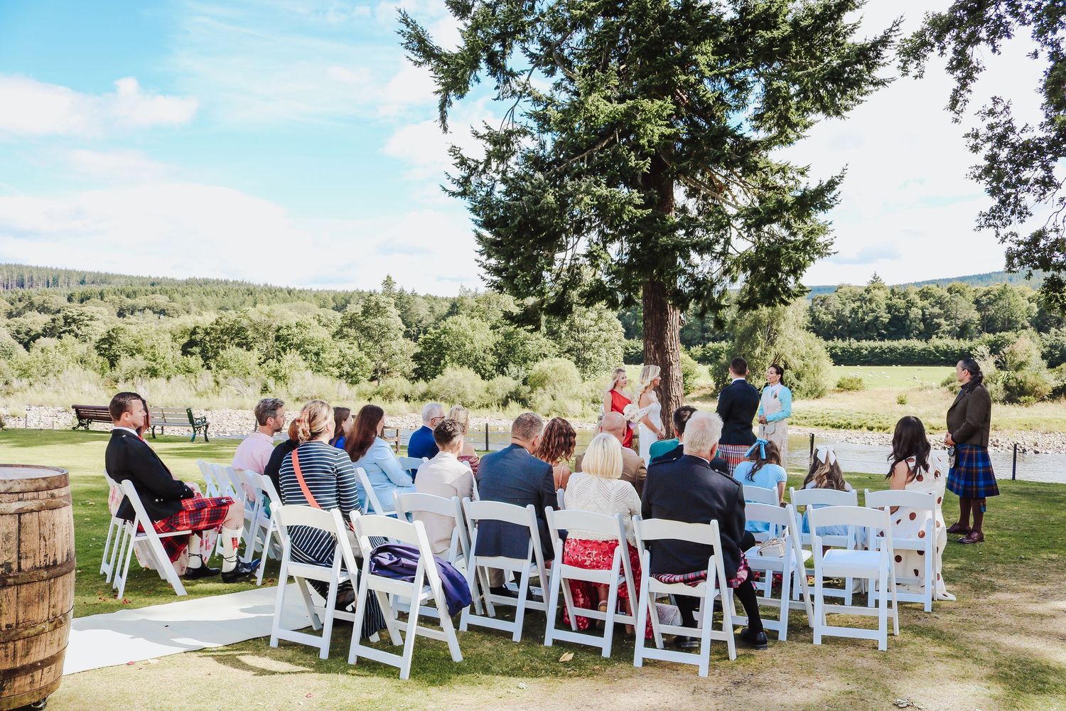 Outdoor wedding ceremony setup with white chairs arranged in rows facing a wooden arch under a large tree.