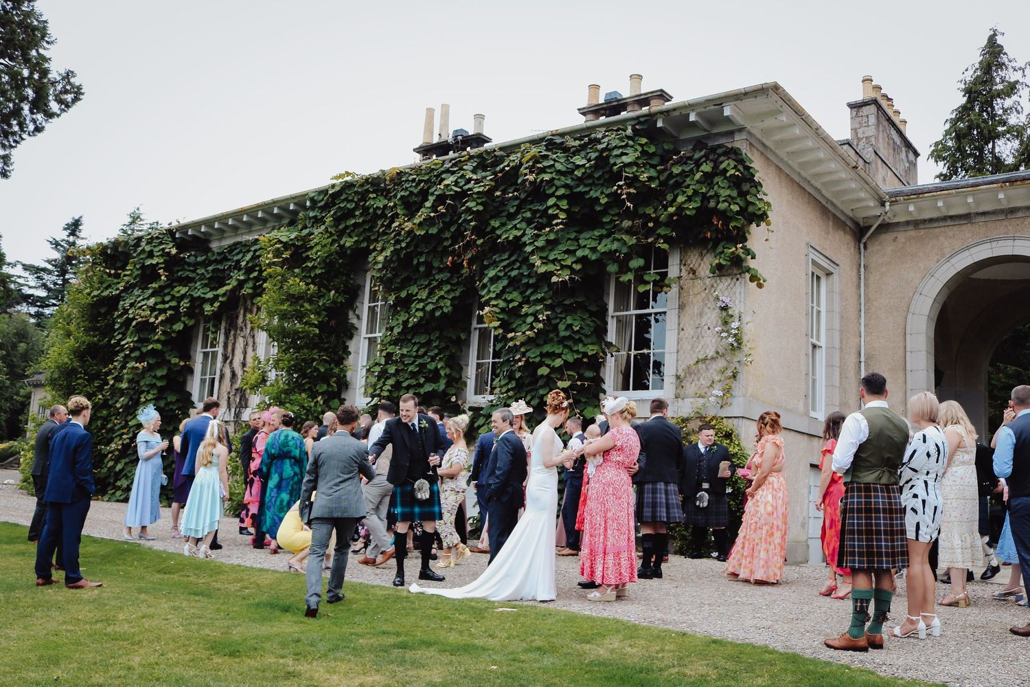 Wedding guests gather outside a historic manor house during an outdoor reception celebration.
