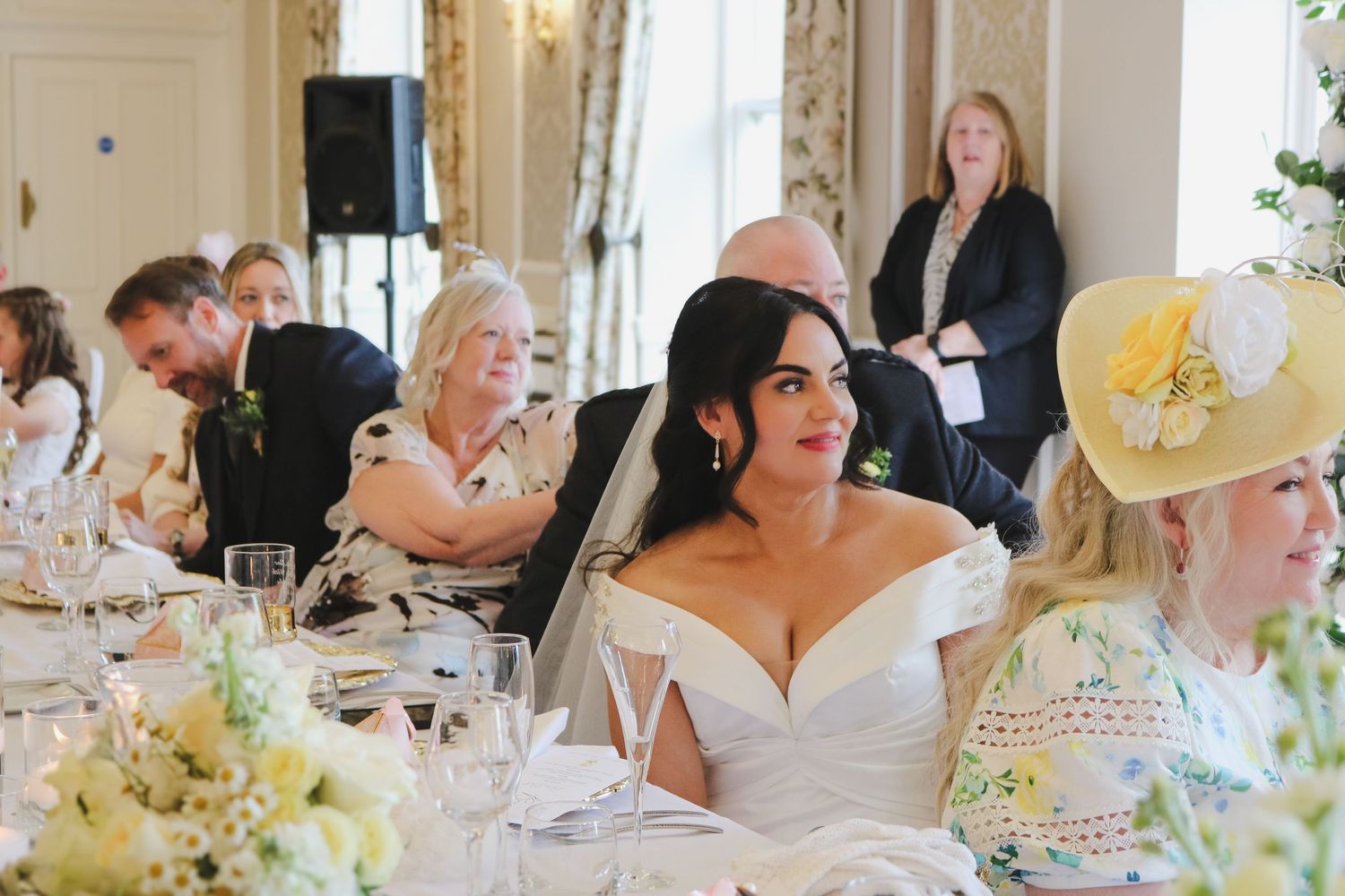 Bride in off-shoulder wedding gown seated at elegant reception table with guests during wedding speeches.