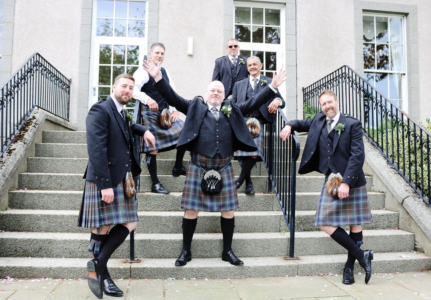 Groom and groomsmen in traditional Scottish kilts posing on stone steps outside a building at a wedding.