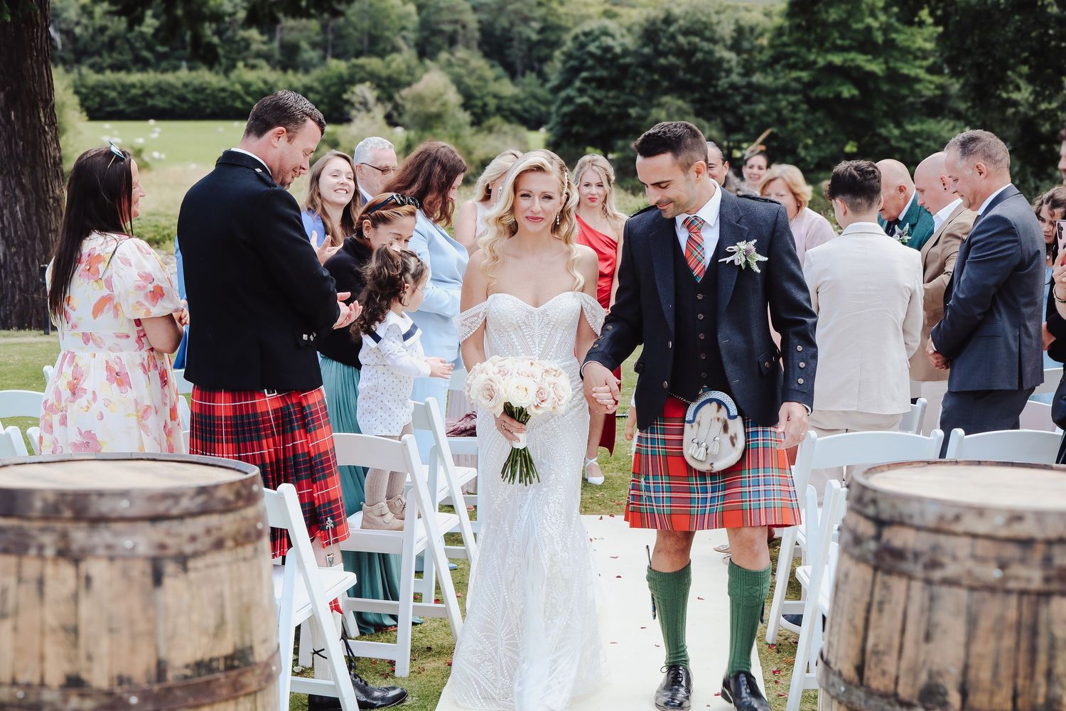 A wedding ceremony outdoors with traditional Scottish kilts and white decor between rustic wooden barrels.