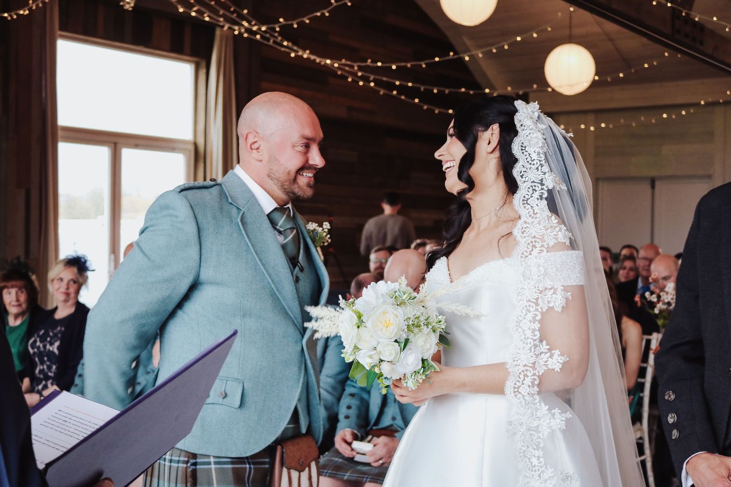 Wedding guests dance and celebrate under twinkling string lights at an indoor reception venue.