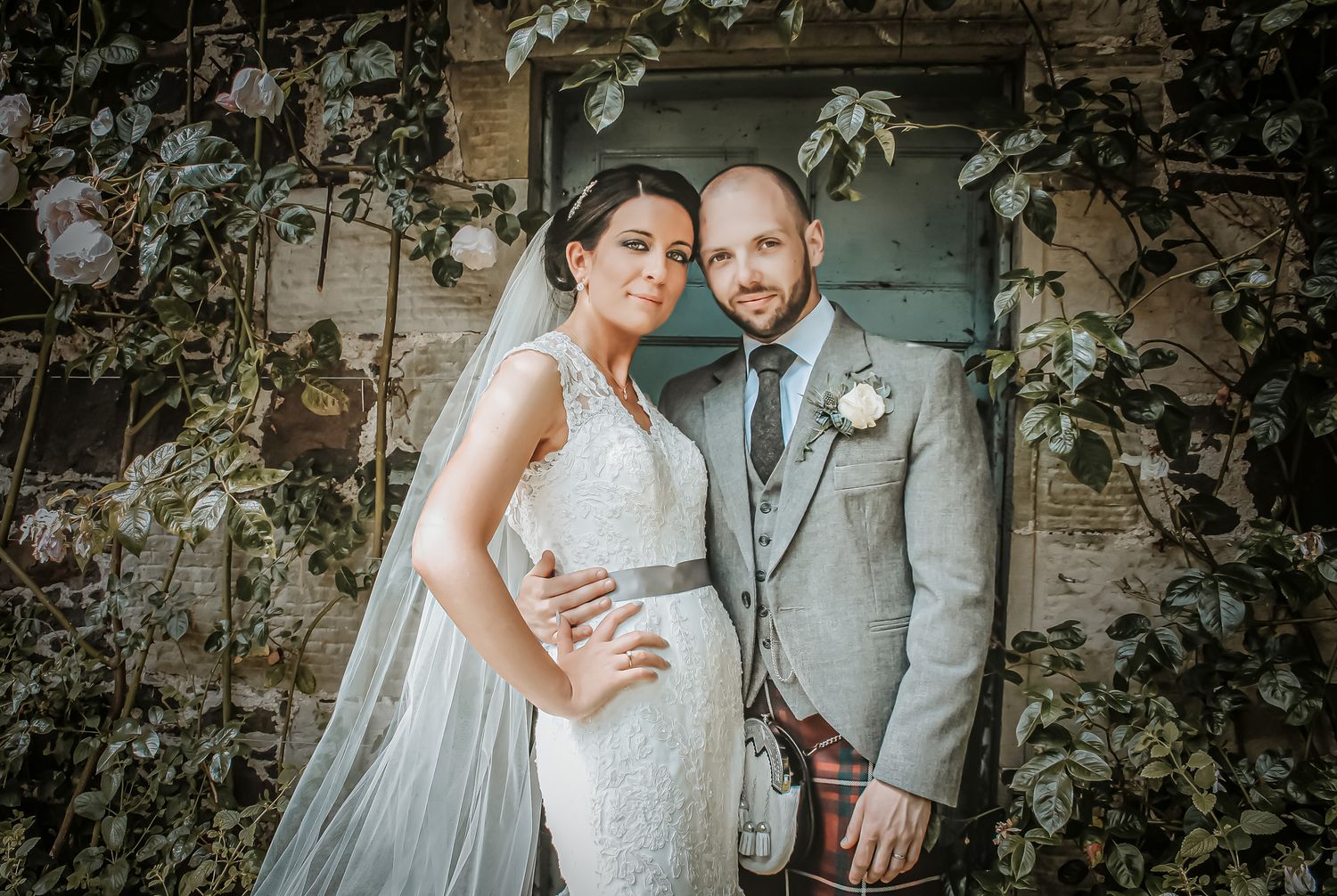 A romantic wedding couple poses against a rustic stone wall covered in climbing vines and white roses.