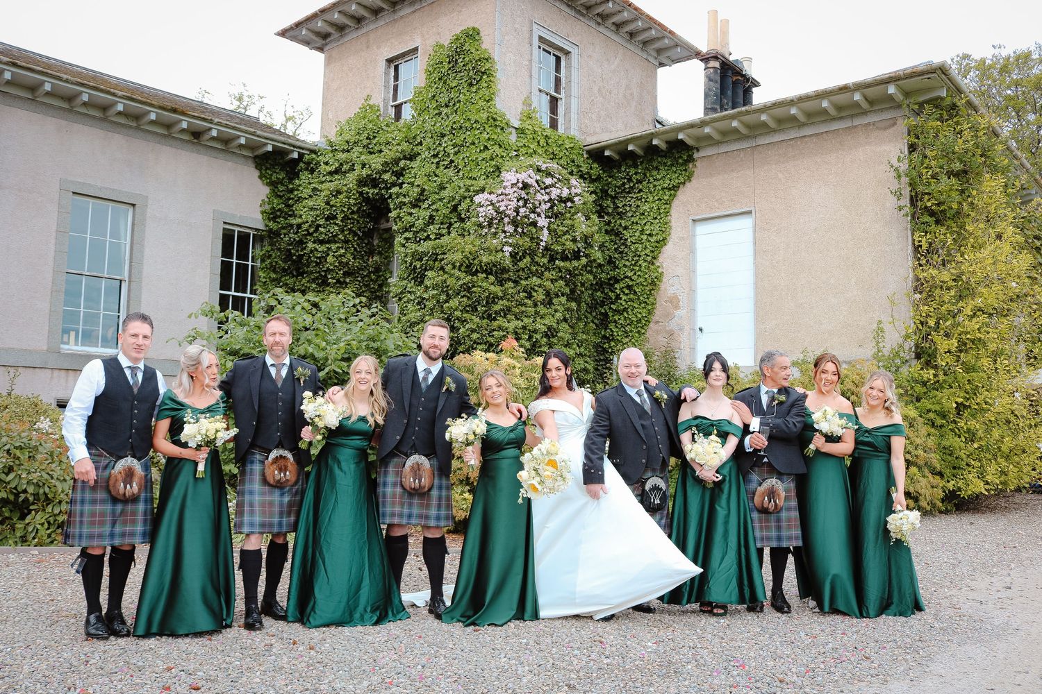 Wedding party in green gowns and kilts poses outside ivy-covered Scottish venue with bride in white gown.