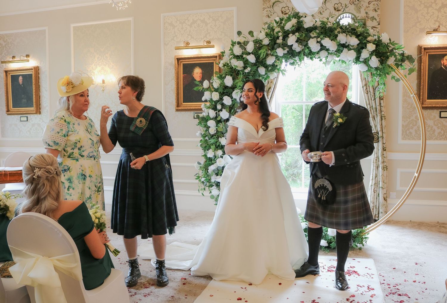 Bride in white gown stands under floral arch with guests in Scottish wedding ceremony venue.