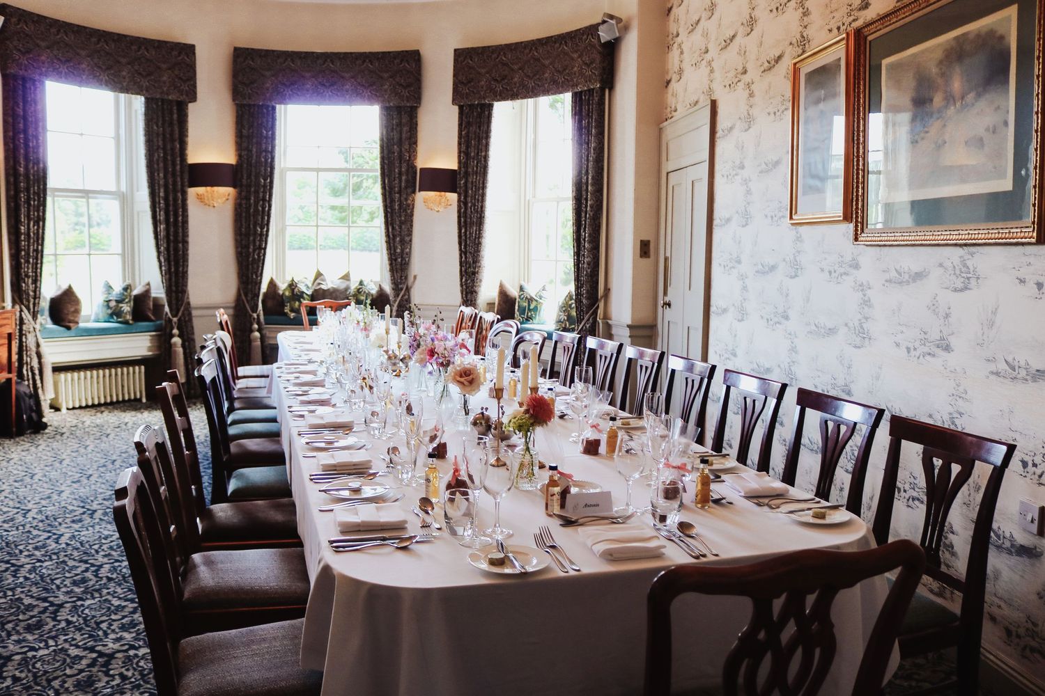 Elegant wedding reception table setting with white linens and floral centerpieces in a stately room with bay windows.