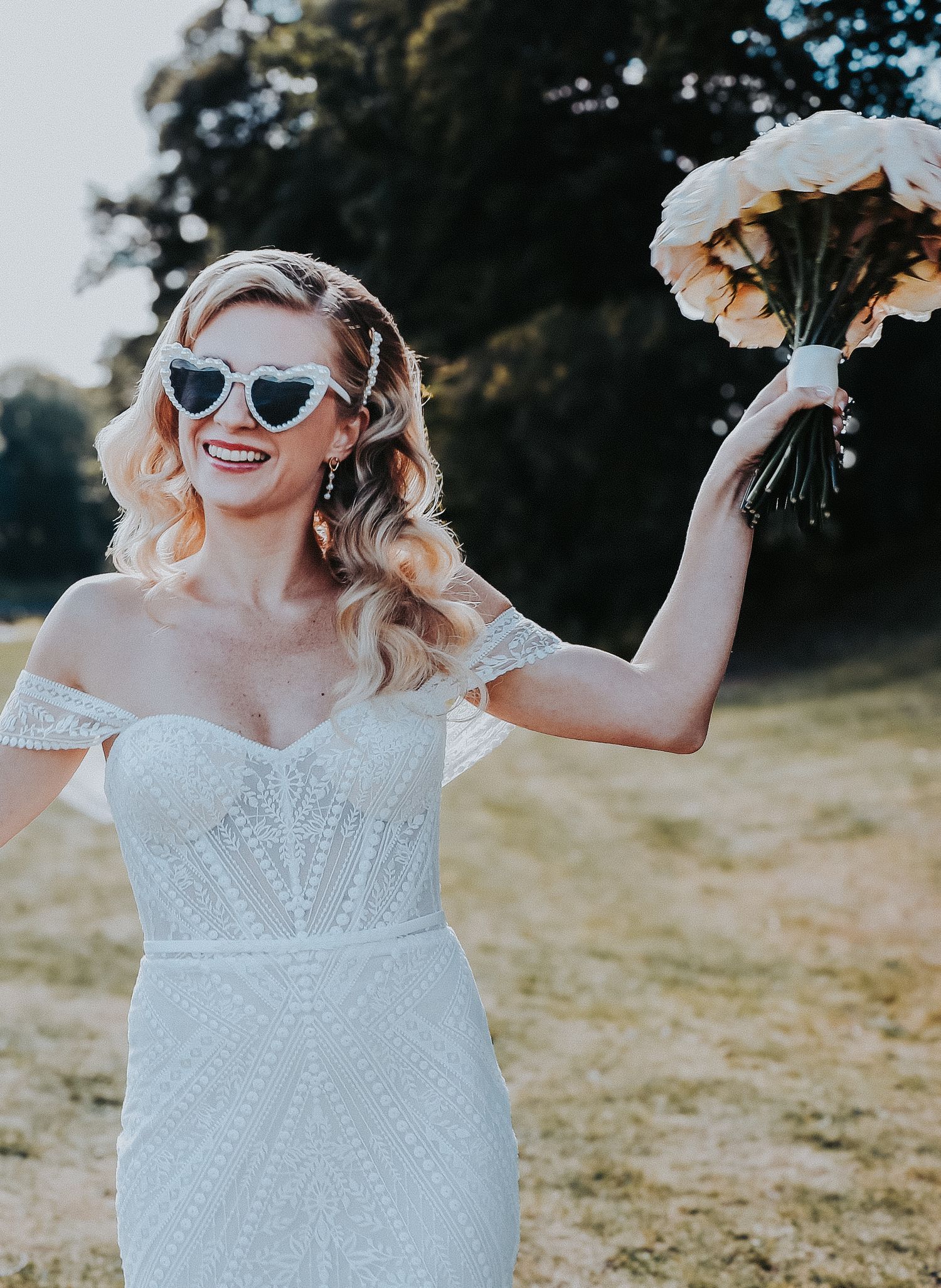 A joyful bride in a white dress and heart-shaped sunglasses poses with flowers outdoors.