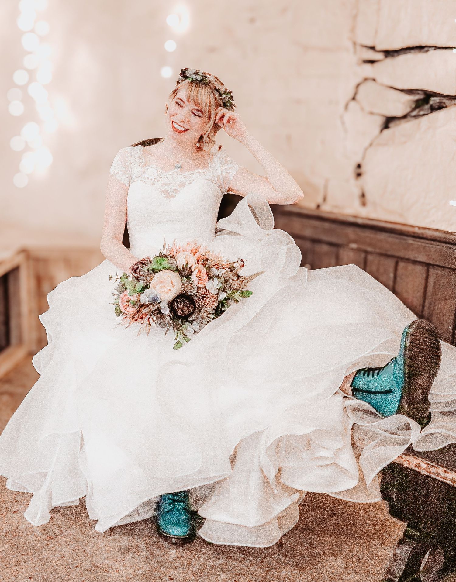 A bride in a flowing white lace gown sits on rustic wooden steps wearing turquoise boots with her wedding bouquet.