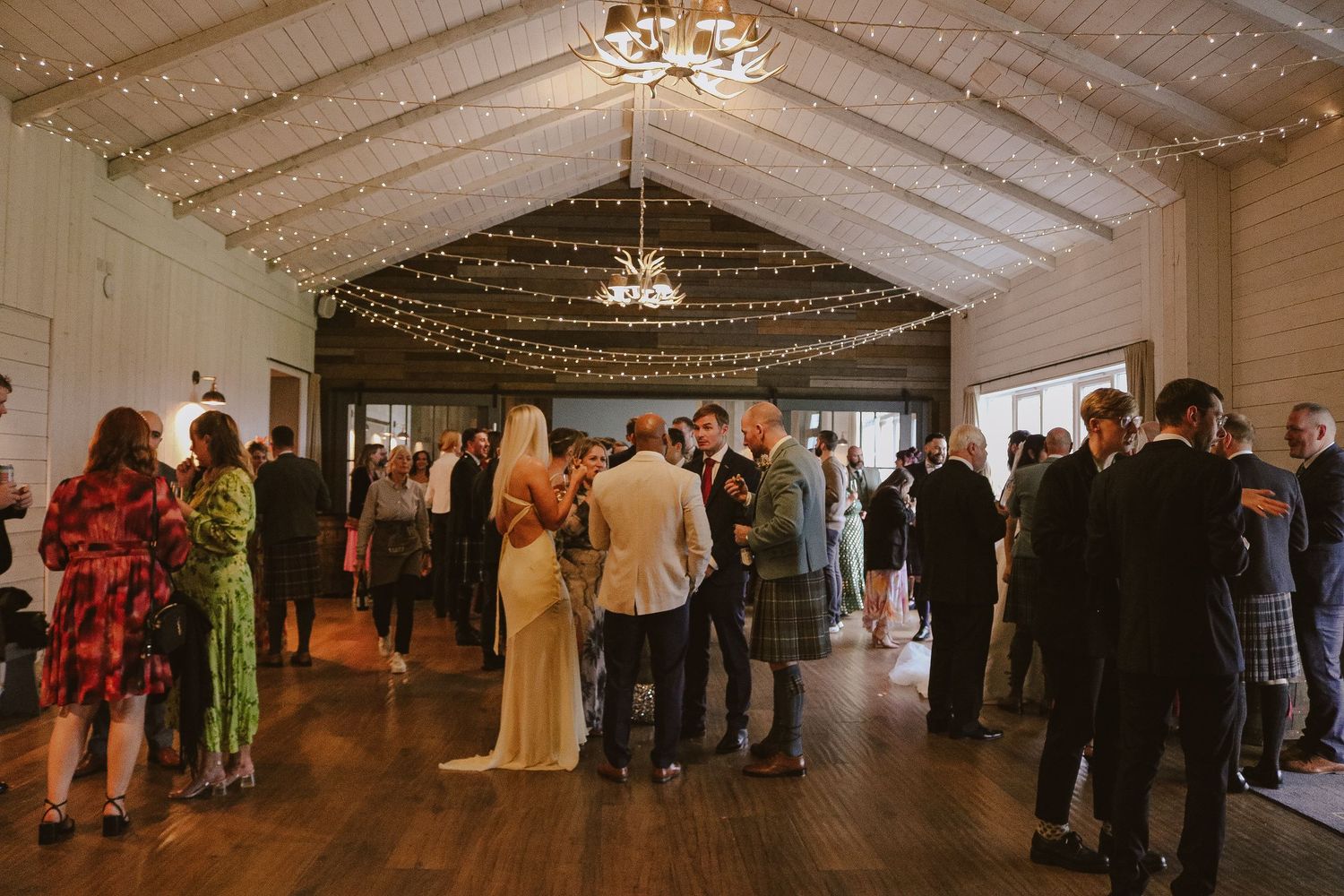 Wedding guests mingle beneath string lights and chandeliers in an elegant white reception hall with vaulted ceilings.