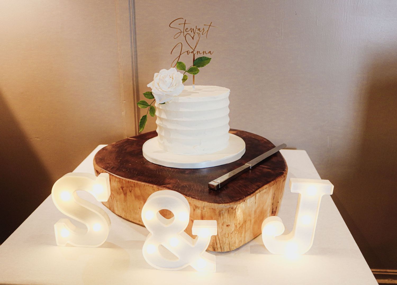 Elegant single-tier white wedding cake decorated with floral accents sits atop rustic wood slice with glowing ampersand sign.