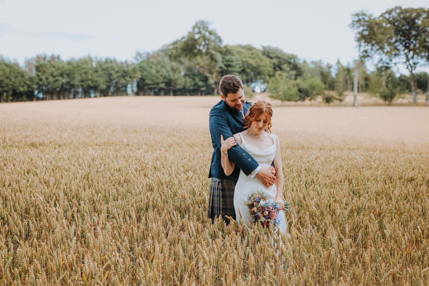 A couple stands embracing in a golden wheat field with trees in the background on a sunny day.