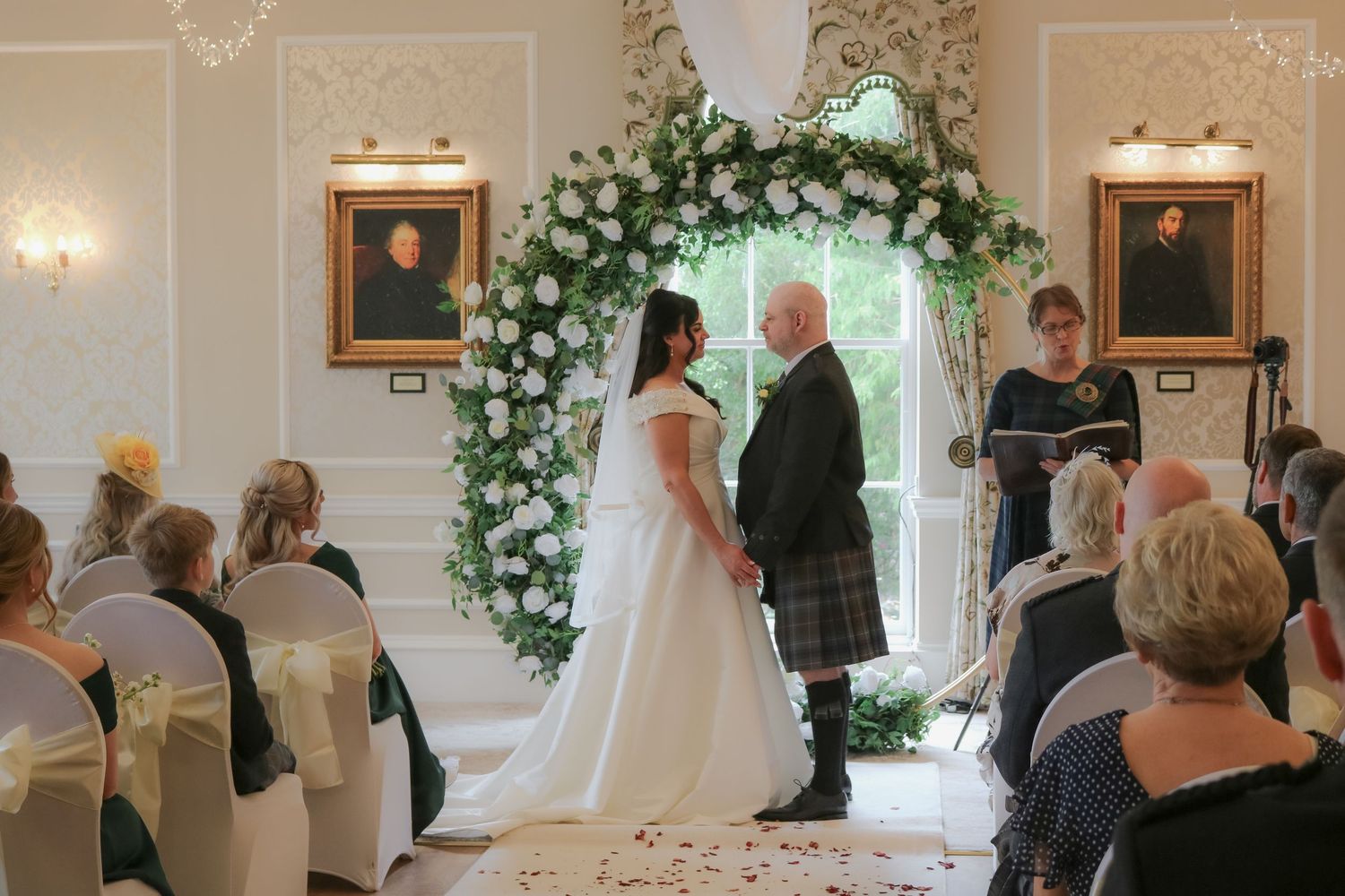 Bride and groom exchange vows under floral arch in elegant white wedding ceremony venue with seated guests.