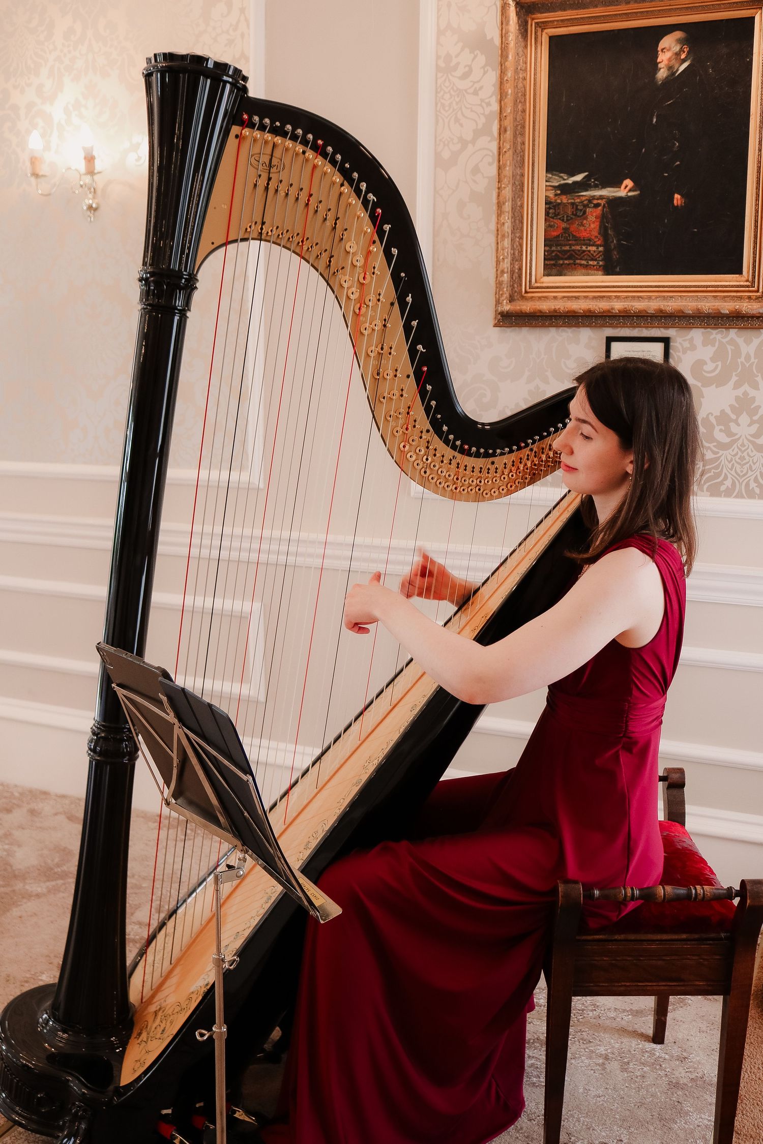 Woman in red gown playing a grand concert harp in an elegant room with classical portrait painting.