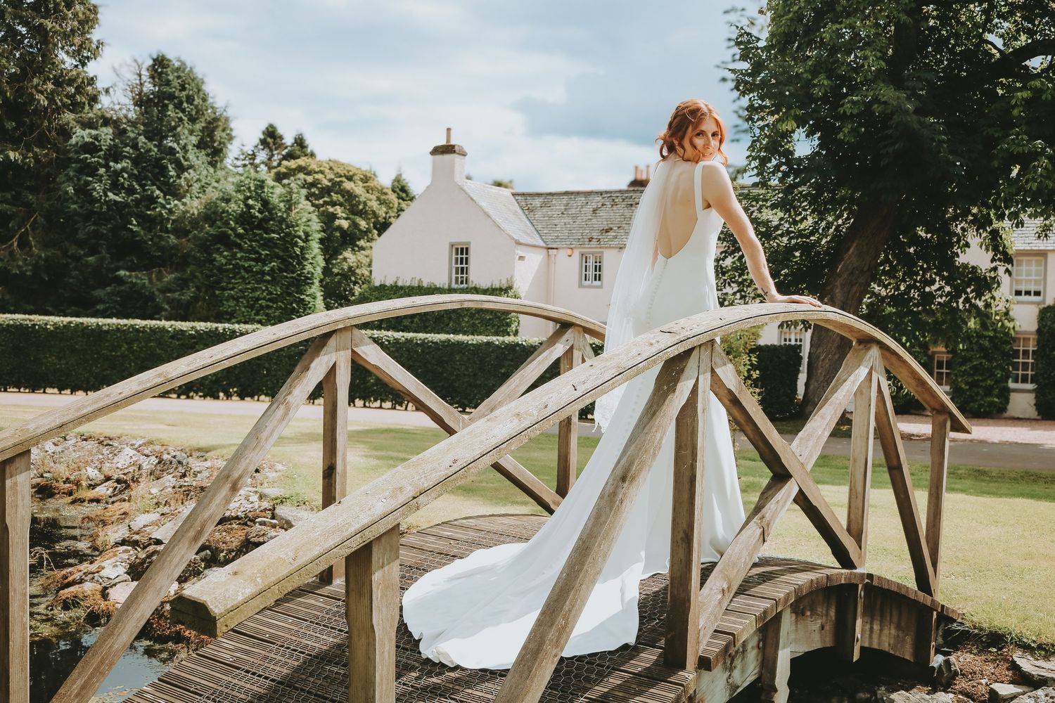 A person in a flowing white wedding dress stands elegantly on a wooden garden bridge in a scenic outdoor setting.