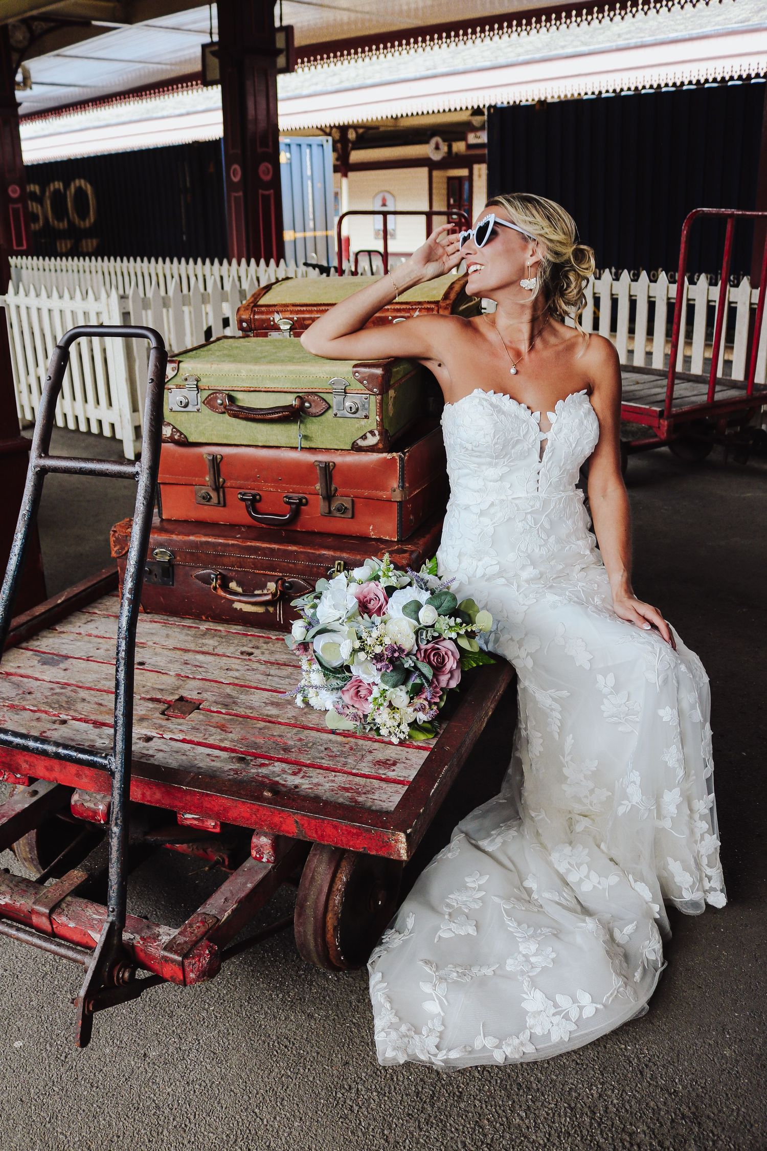 A bride in a white wedding dress poses with vintage luggage on a rustic red cart at a train station platform.