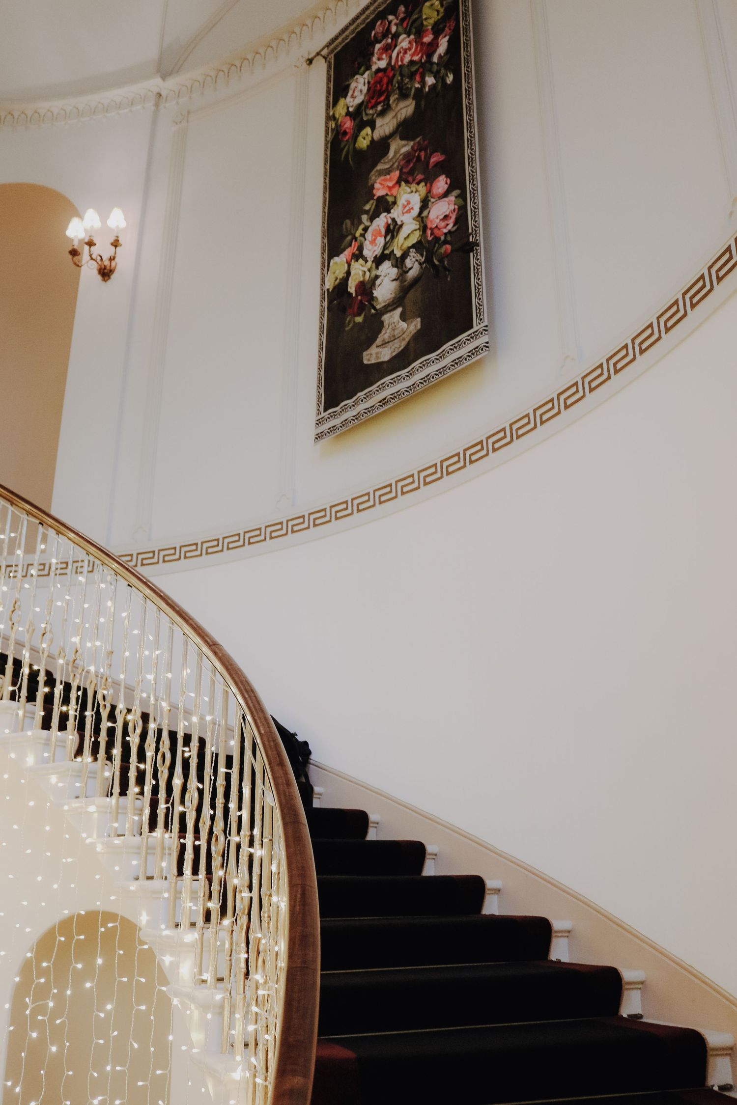 Elegant curved staircase with black carpet and white walls featuring fairy lights and floral artwork above.