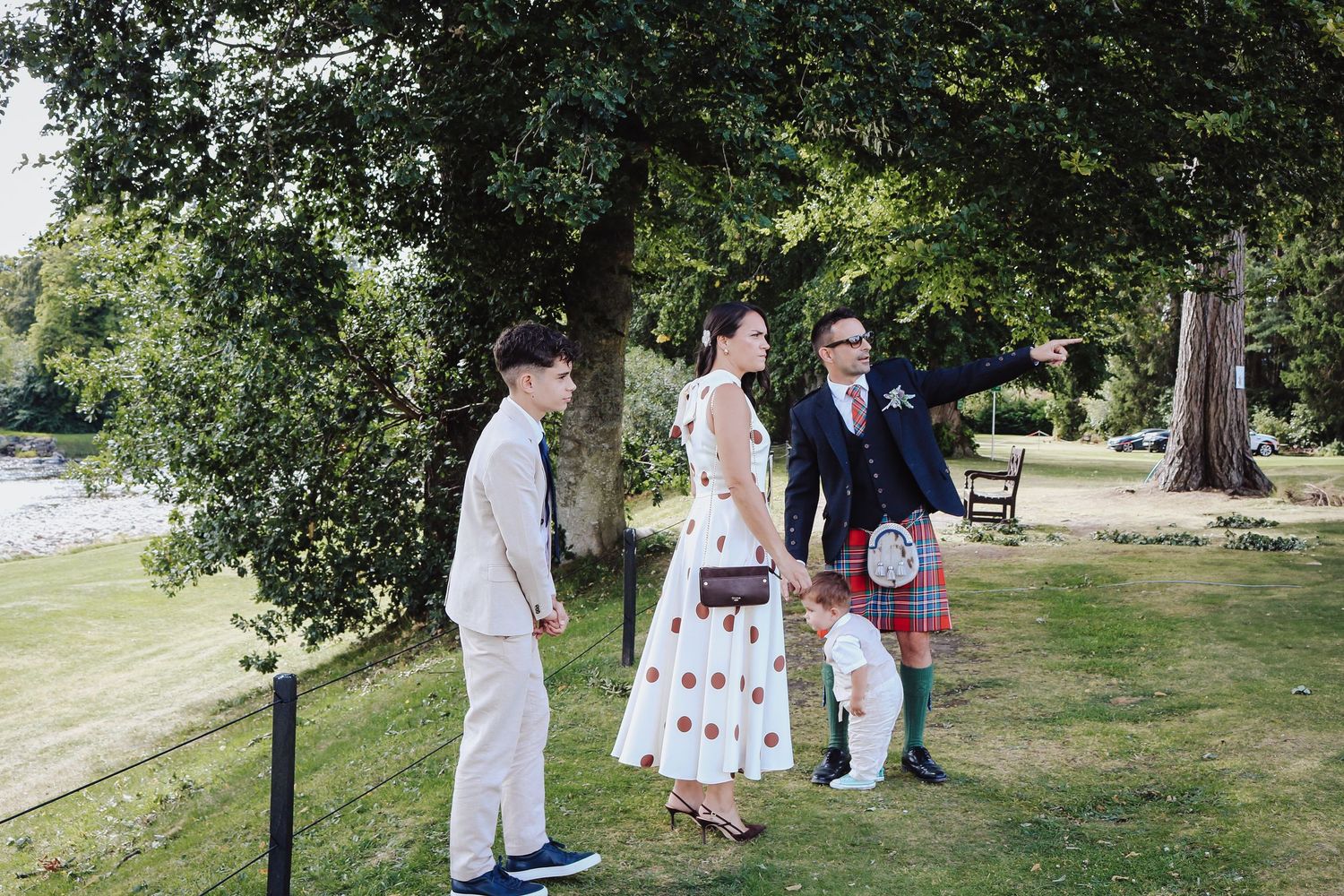 People in traditional Scottish attire and summer clothing gather outdoors on a grassy lawn near trees on a sunny day.