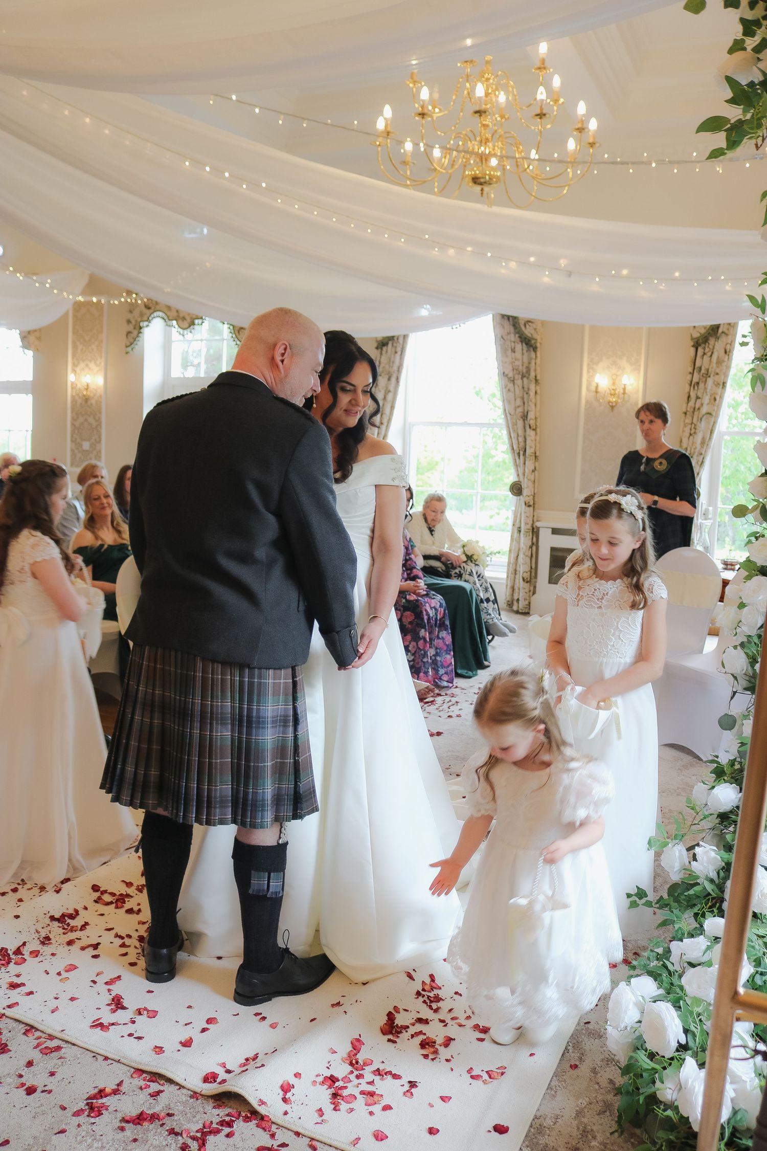 Bride and groom in tartan kilt exchange vows at indoor wedding ceremony with flower girls scattering red rose petals.
