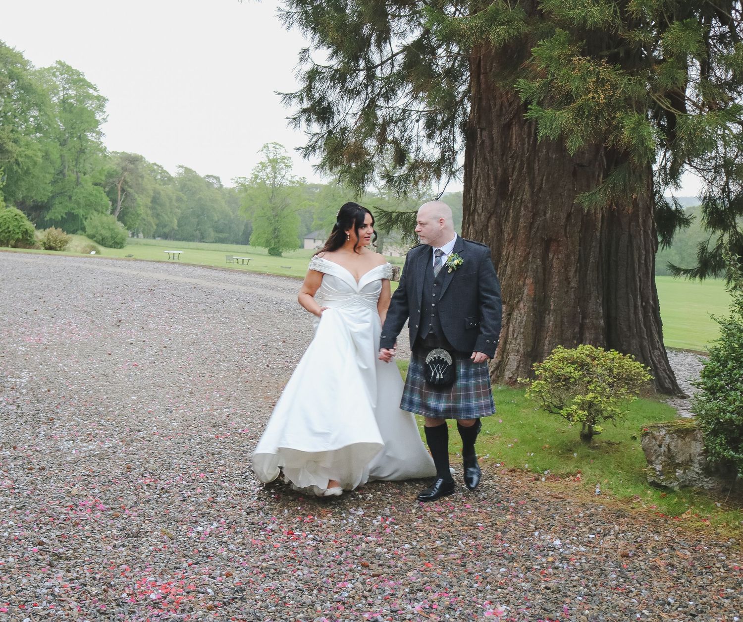 Bride in white gown and groom in Scottish kilt walk hand in hand on gravel path surrounded by lush green trees.