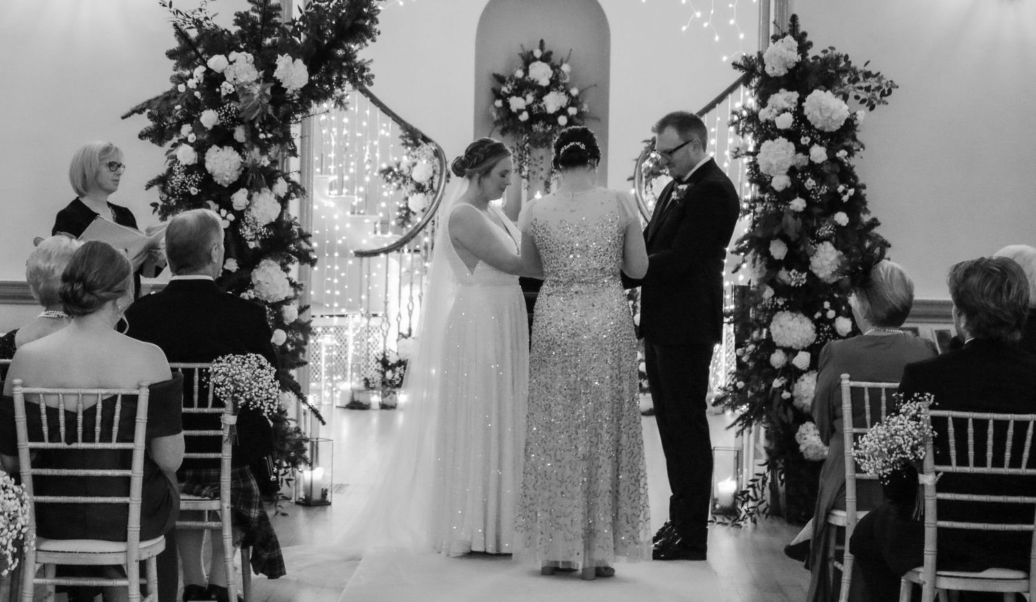 Black and white wedding ceremony photo showing couple at the altar with floral arrangements and guests seated in chairs.