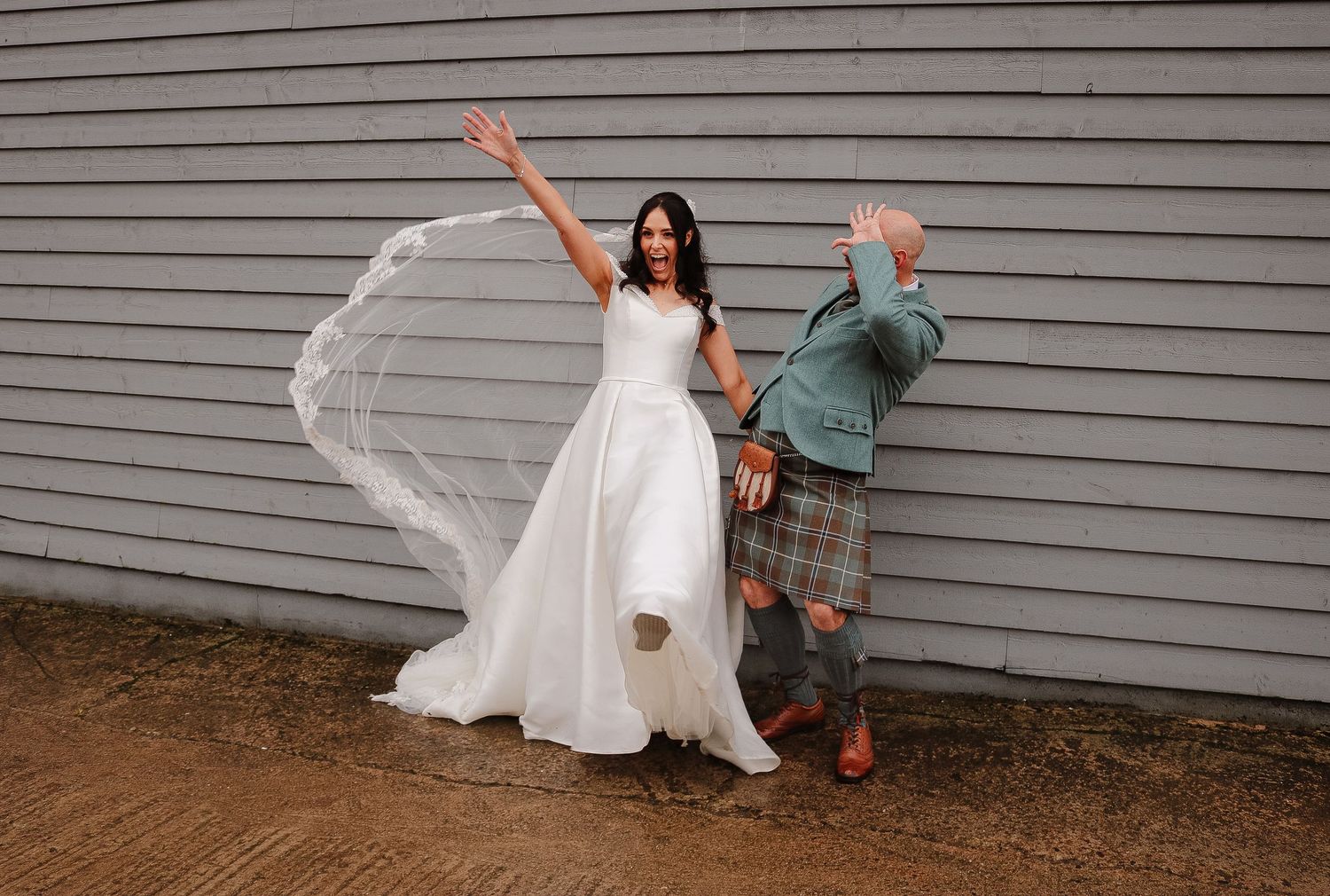 A couple shares a playful dance sequence against a white siding wall in wedding attire.