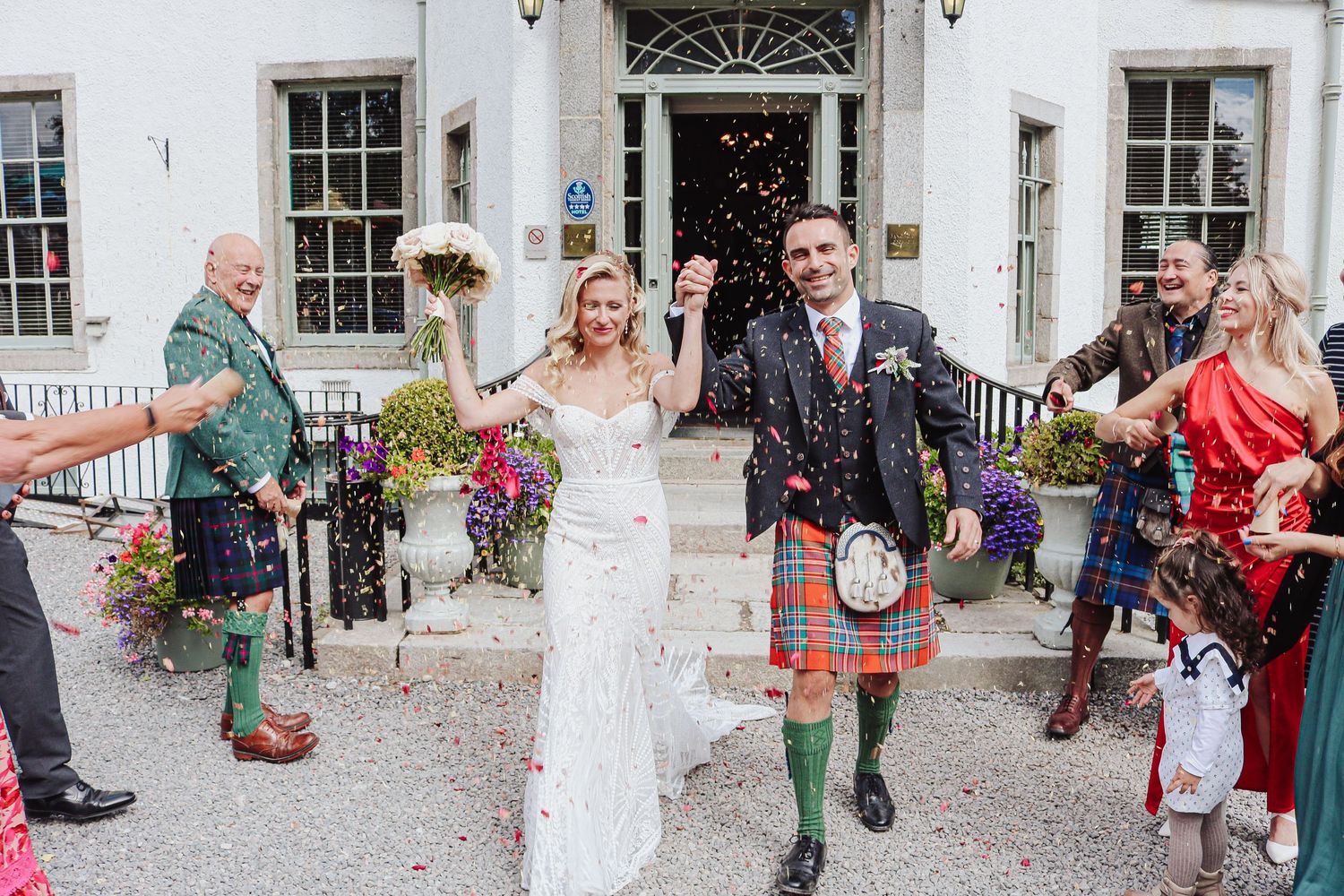 Wedding guests throw confetti over the newlyweds outside a white stone building during a joyous celebration.