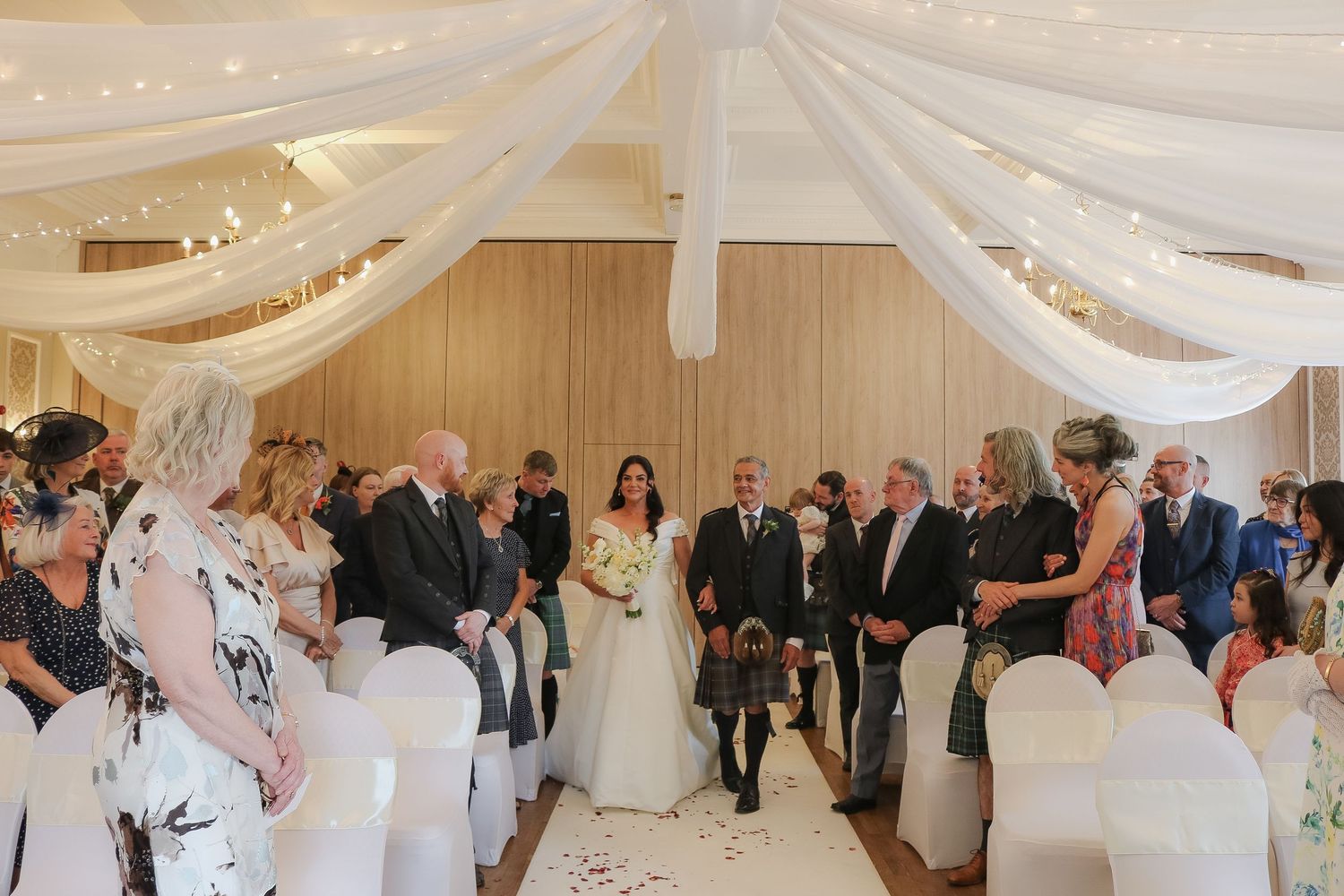 Elegant indoor wedding ceremony with draped white fabric ceiling, guests seated in white chairs, and bride walking down the aisle.