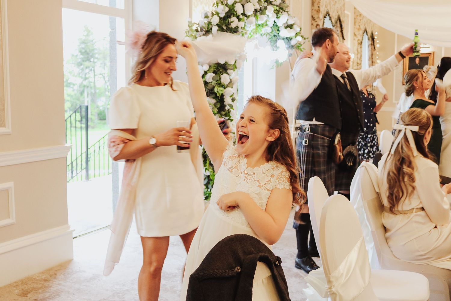Joyful girl laughing at a wedding reception while guests celebrate in an elegant white decorated venue.