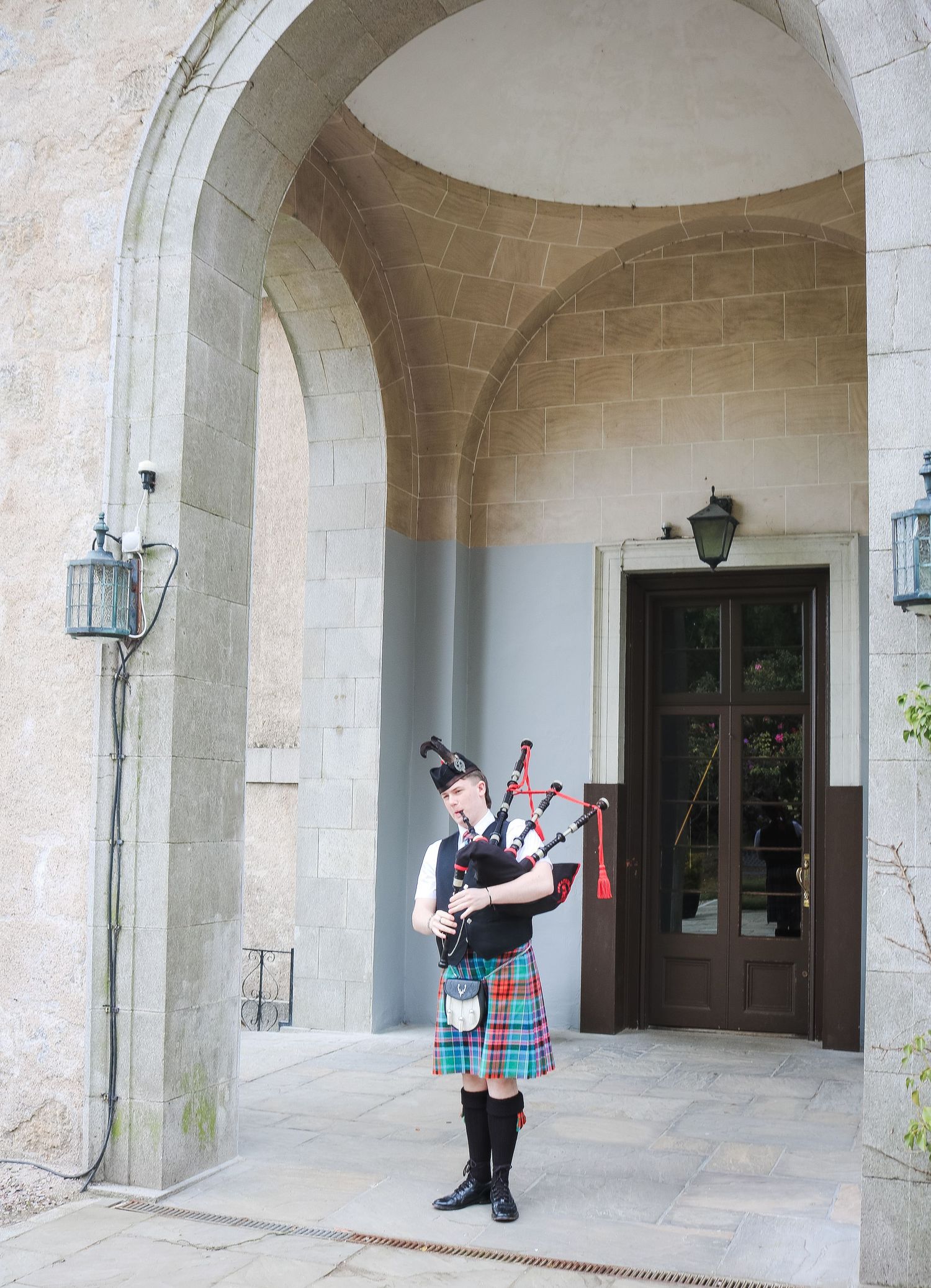 A bagpiper in traditional Scottish tartan kilt performs under a stone archway at an elegant venue entrance.