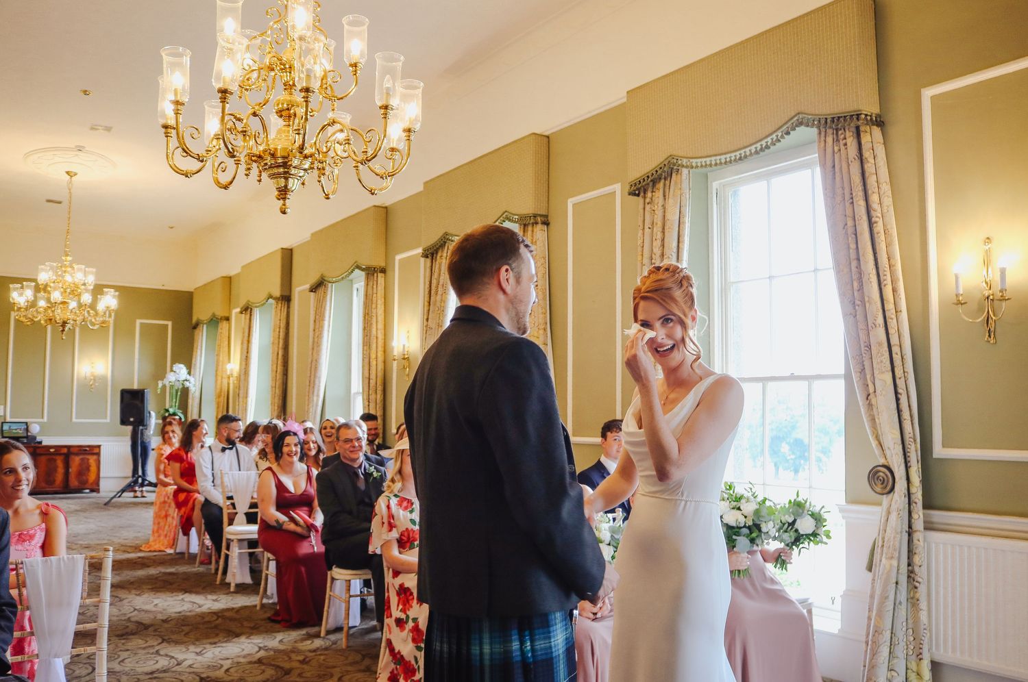 Elegant wedding ceremony takes place in a grand ballroom with crystal chandeliers and sage green walls.
