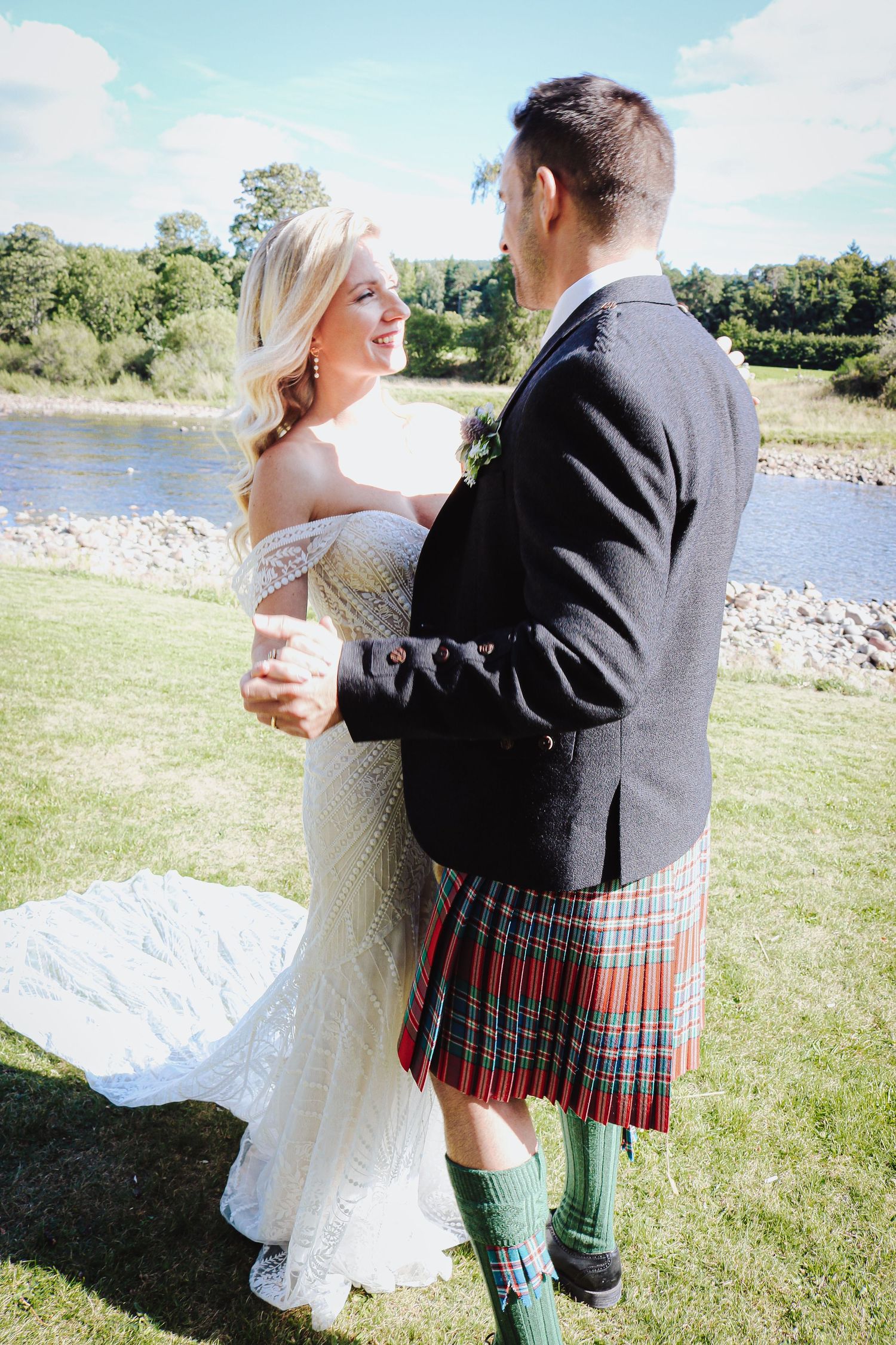 A couple in wedding attire by a river, with the groom wearing traditional Scottish kilt and green socks on a sunny day.