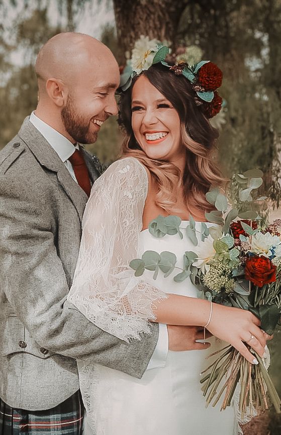 A wedding couple shares a joyful moment outdoors, with the bride wearing a floral crown and holding a bouquet.
