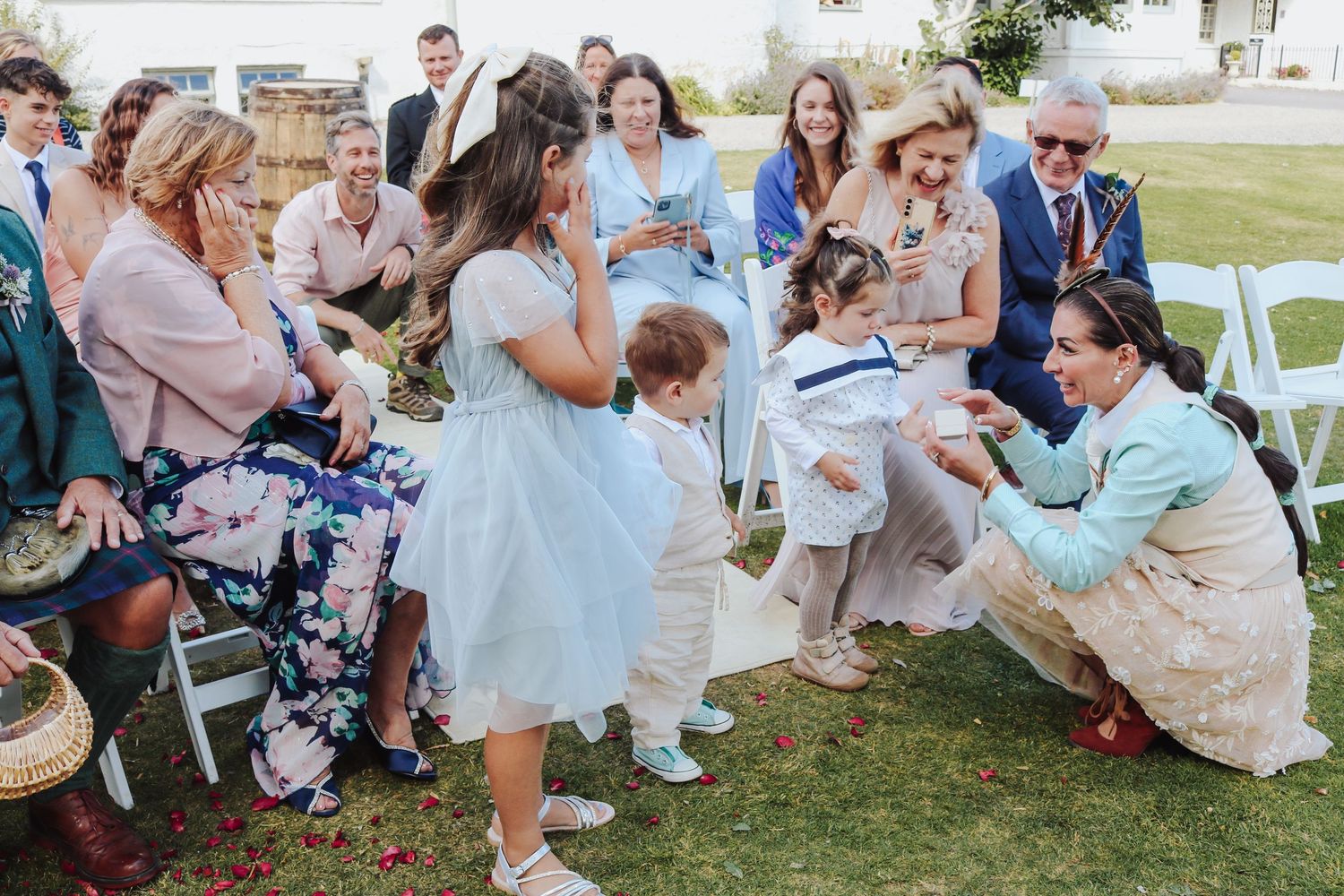 A bending figure in light blue proposes to a person in a blue dress at an outdoor wedding ceremony while guests look on.