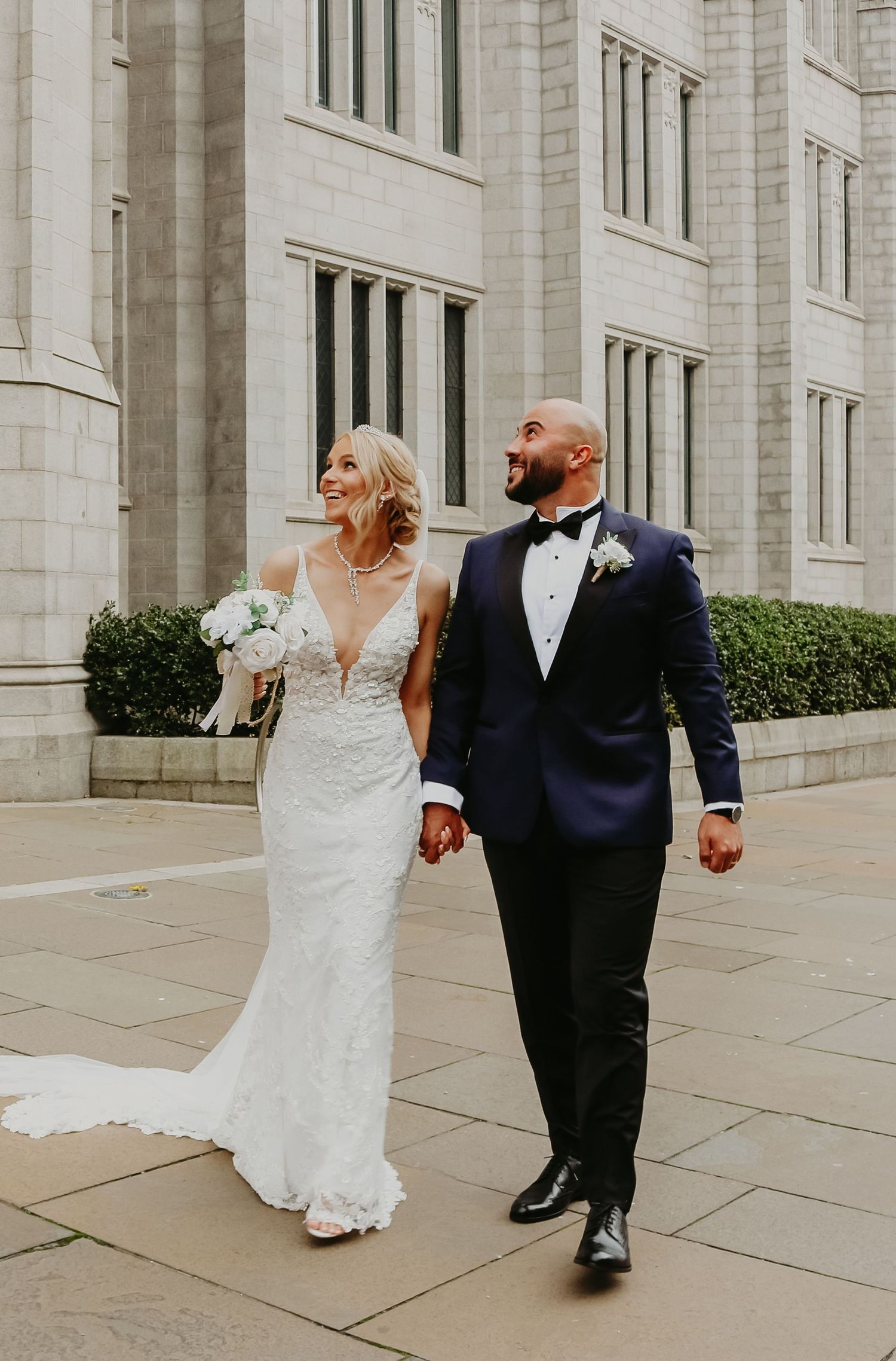 A couple walks hand in hand along a grand stone building exterior in wedding attire.