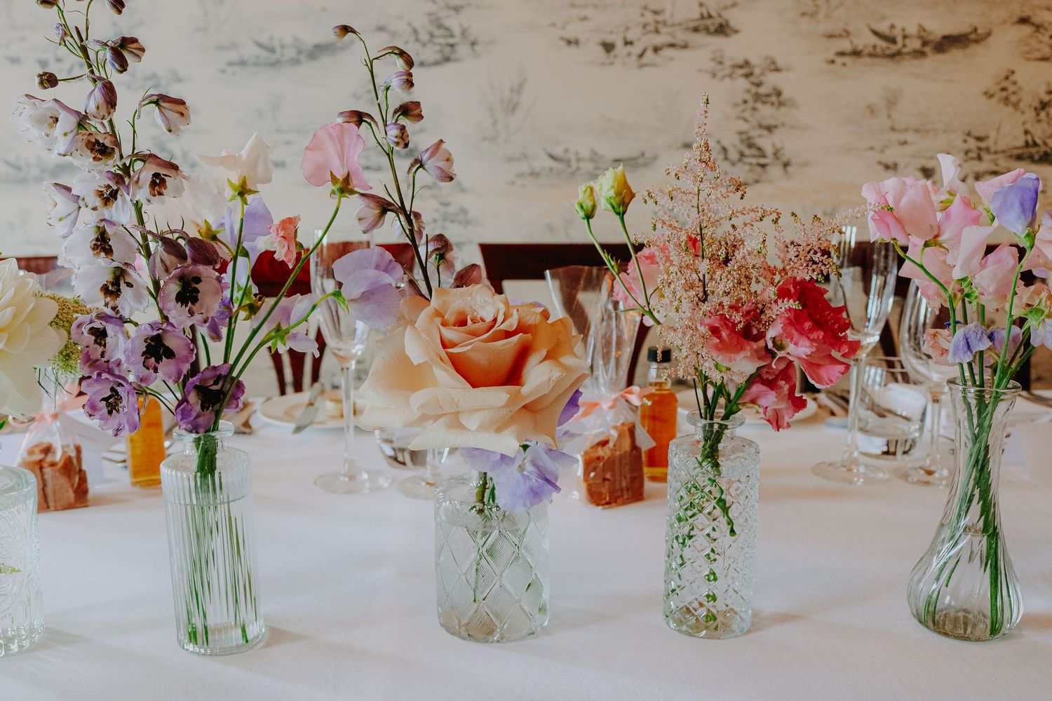 Delicate floral arrangements in glass vases featuring pink, peach and purple blooms against a textured wall backdrop.