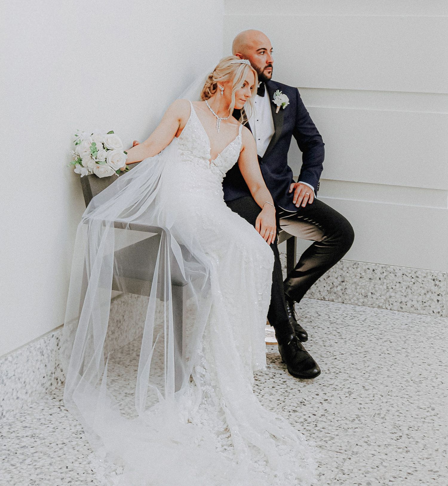 A bride in a fitted lace gown with long veil poses with her groom against a white wall on their wedding day.
