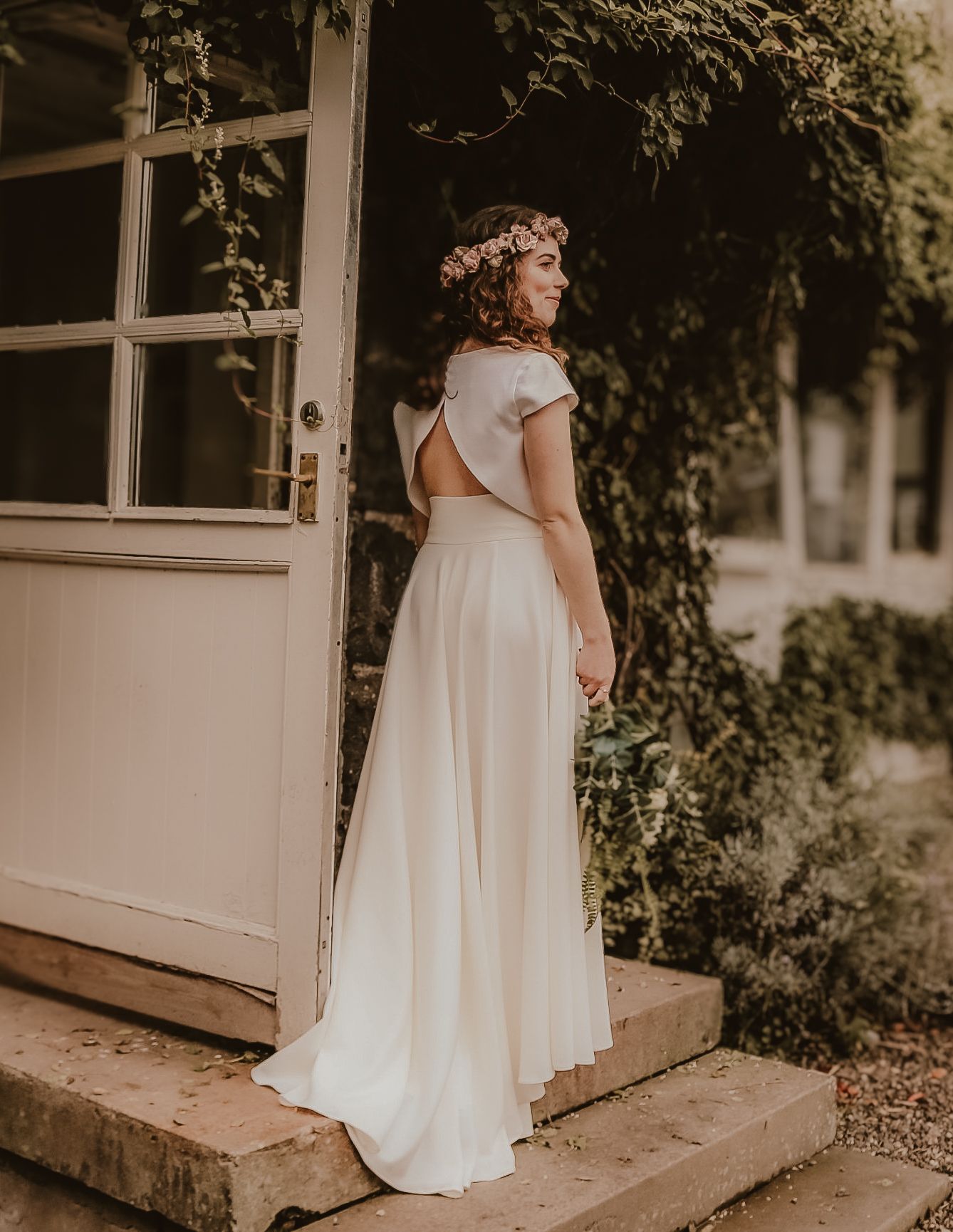 A bride in a flowing two-piece wedding dress with cap sleeves stands on stone steps near a white door with climbing vines.